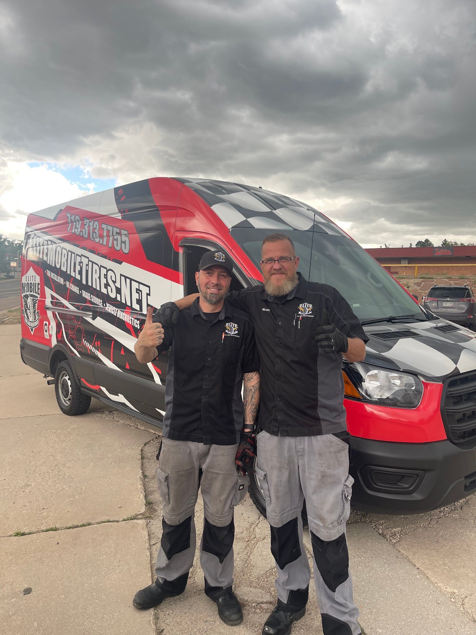 Two men stand next to a checkered flag-themed van. One gives a thumbs up. Overcast sky. | Elite Mobile Tires and Maintenance