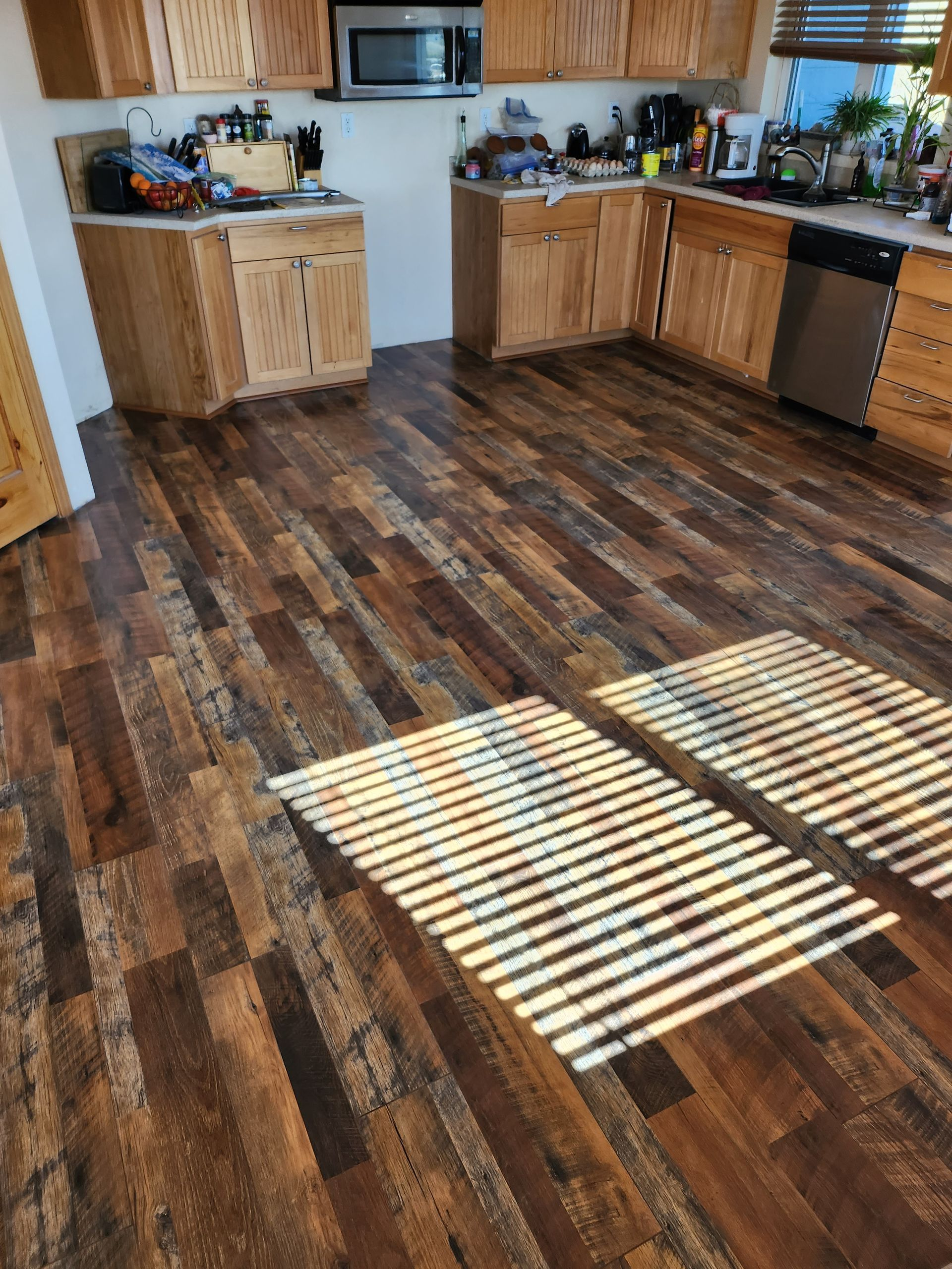 Kitchen with wood-look flooring and light wood cabinets. Sunlight streams across the floor from a window.