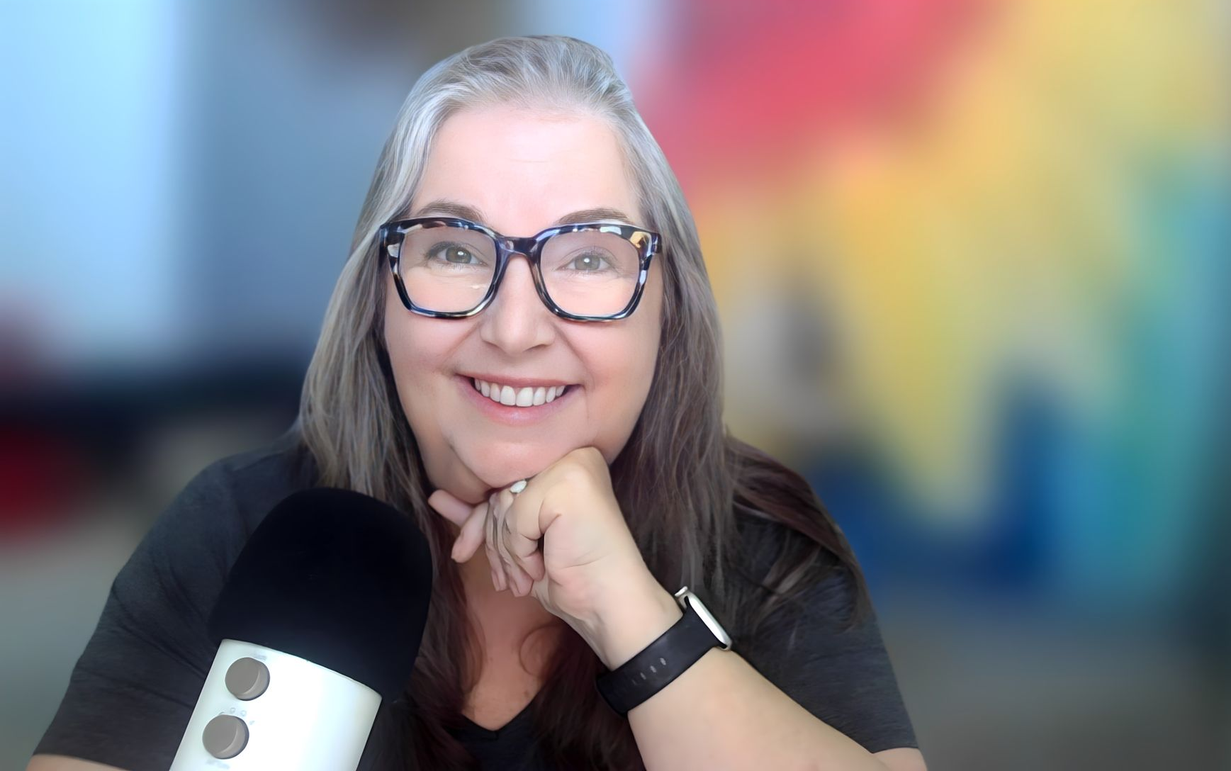 Woman with glasses smiling, hand on chin next to a microphone, colorful background.