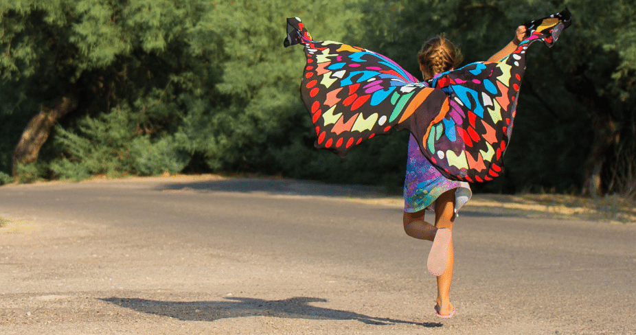 Child in colorful butterfly wings running down a road with arms outstretched.