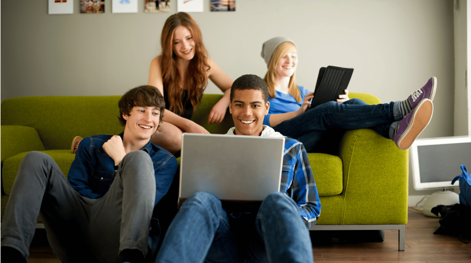 Four young adults sit and recline on and around a green couch, using a laptop and tablet, smiling.