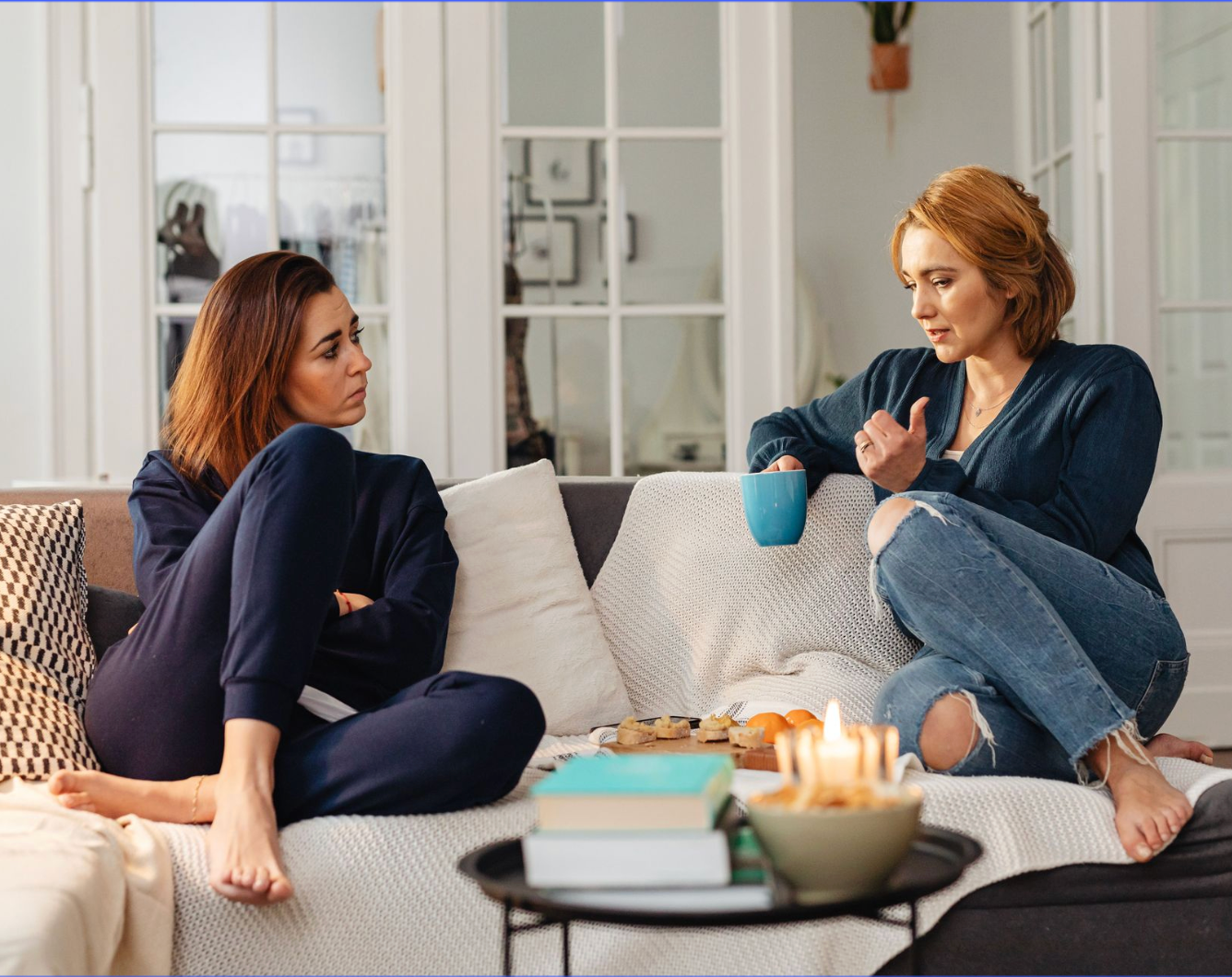 Two women talking on a couch in a cozy living room with coffee mugs and candles