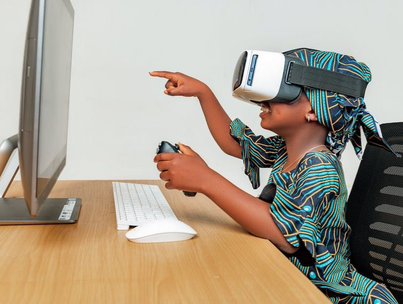 Child wearing a VR headset points at a computer monitor while using a mouse at a desk