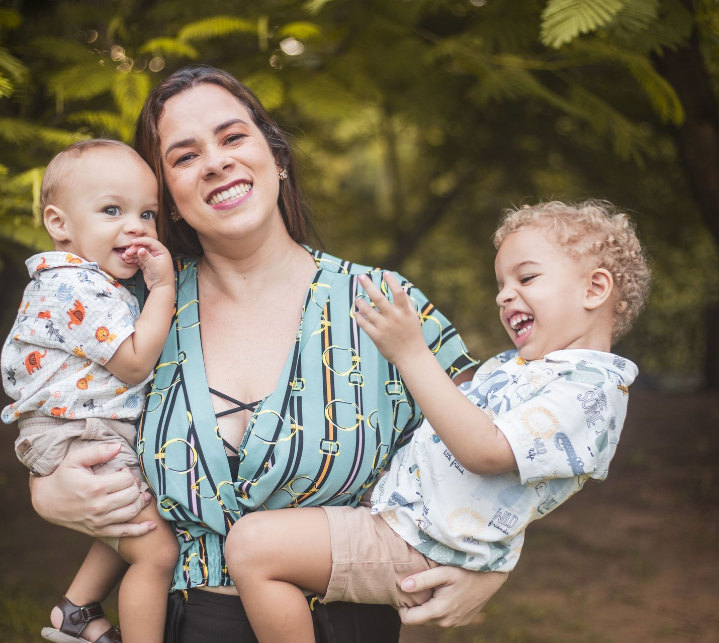Woman holding two smiling toddlers in a leafy outdoor setting