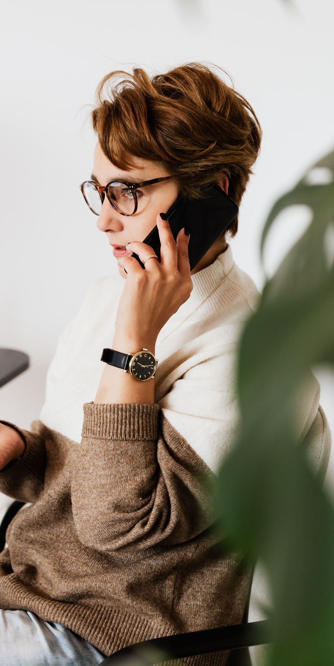 Person with glasses and short hair talks on a phone while sitting indoors in a beige sweater.