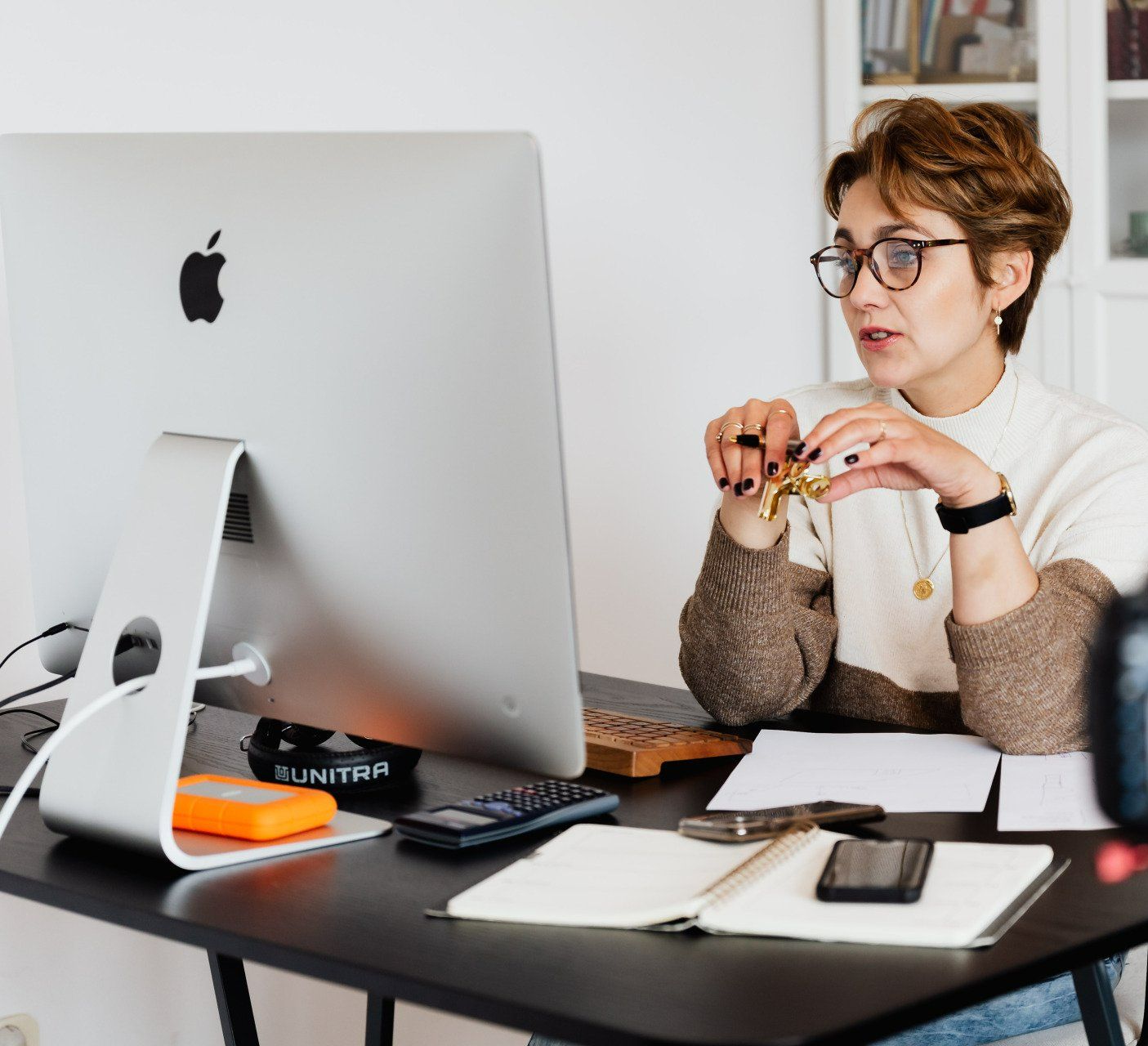 Woman at desk with computer, looking at the screen, talking and holding objects; papers and phone visible.