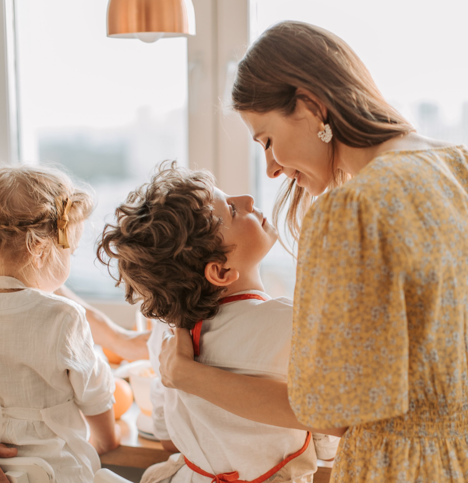 Woman embraces a child in a kitchen, looking lovingly into his eyes. A second child is nearby. Yellow floral dress.