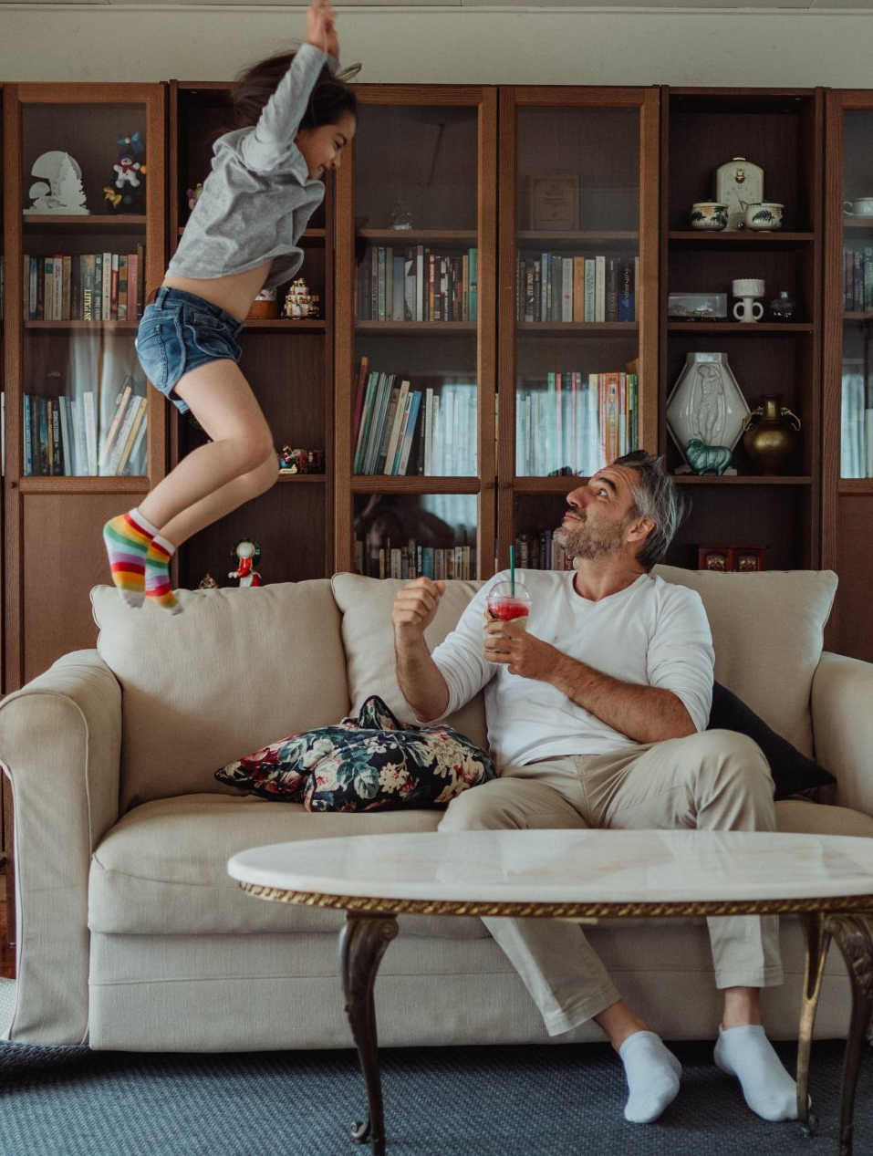 Girl jumping near a man on a couch. Living room with bookshelves in the background. Man holding a drink.