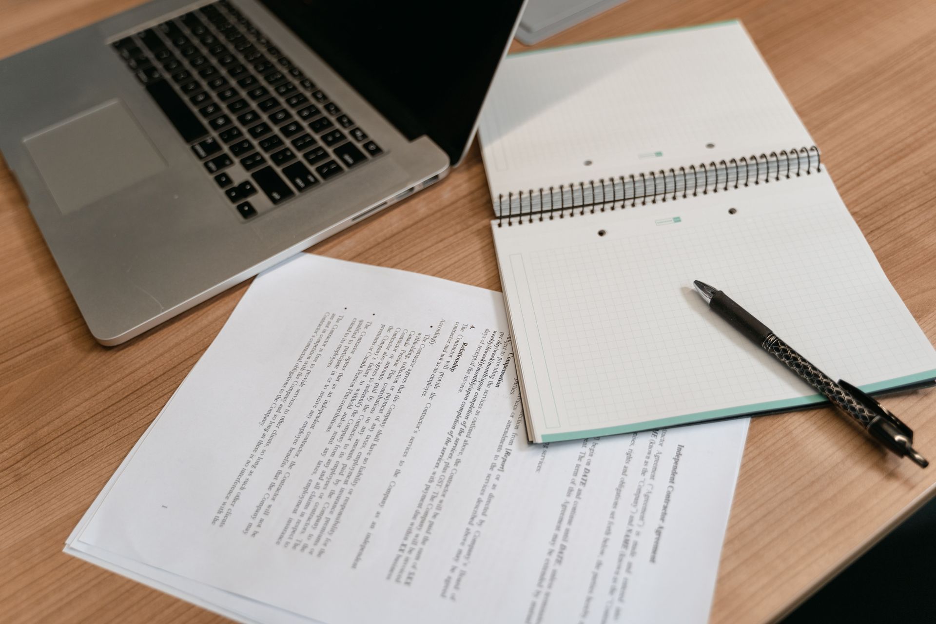 Laptop and notepad with pen on a wooden desk, with printed documents spread nearby