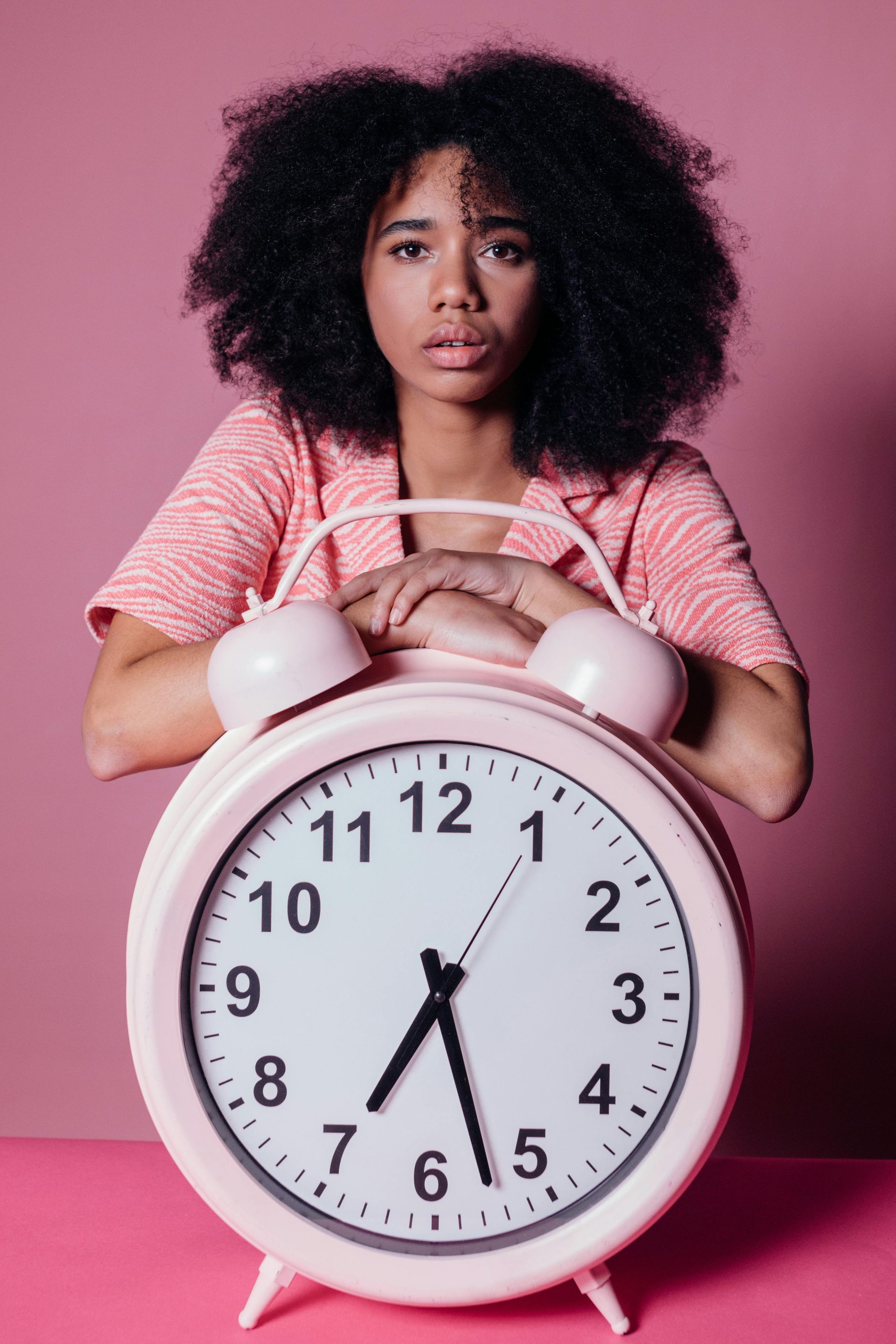 Woman with curly hair, arms on large pink alarm clock, against pink backdrop, looking concerned.