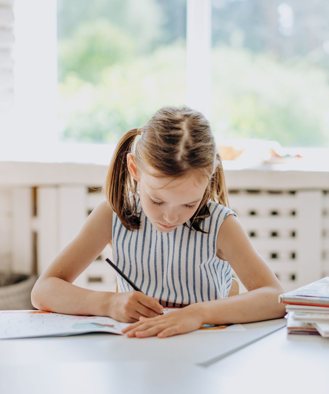 Girl writing in a notebook at a white desk by a bright window.