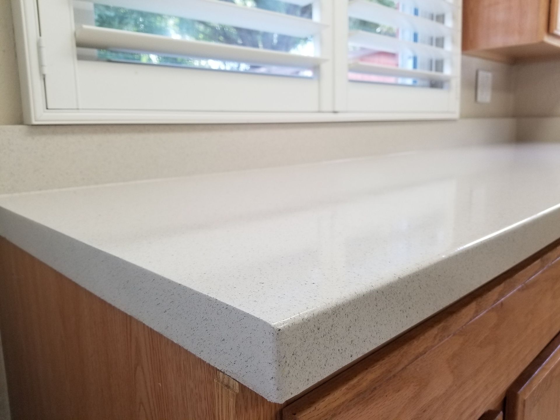 A close-up of a smooth, speckled off-white quartz countertop with a square edge, set above wooden kitchen cabinets.
