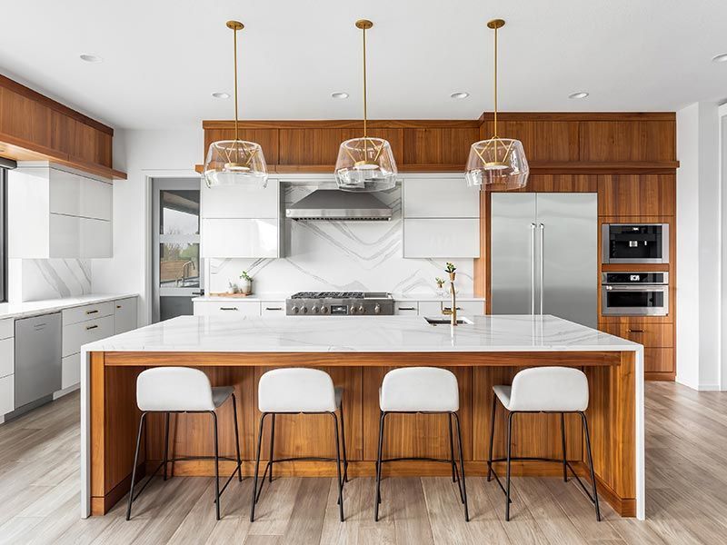 Modern kitchen with wood cabinetry, white countertops, stainless steel appliances, and four white stools at an island.