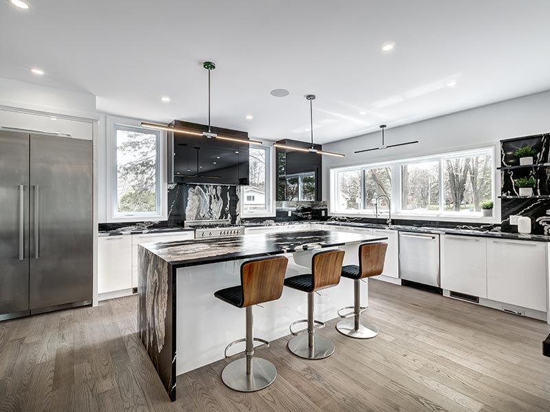 Modern kitchen featuring a white island with three stools, stainless steel appliances, and large windows with light floors.