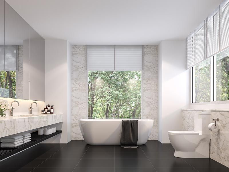 Modern bathroom with a freestanding tub, a marble double vanity, and a toilet against a window with a forest view.