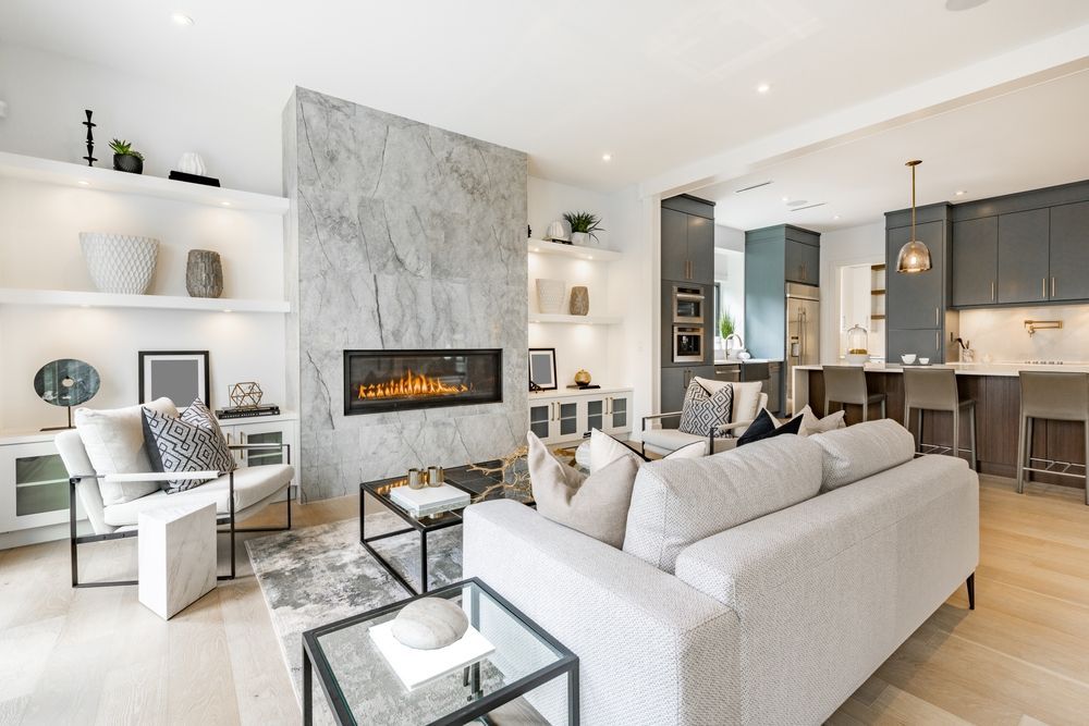 A modern, bright living room with a large stone fireplace, light-colored furniture, and an open kitchen in the background.