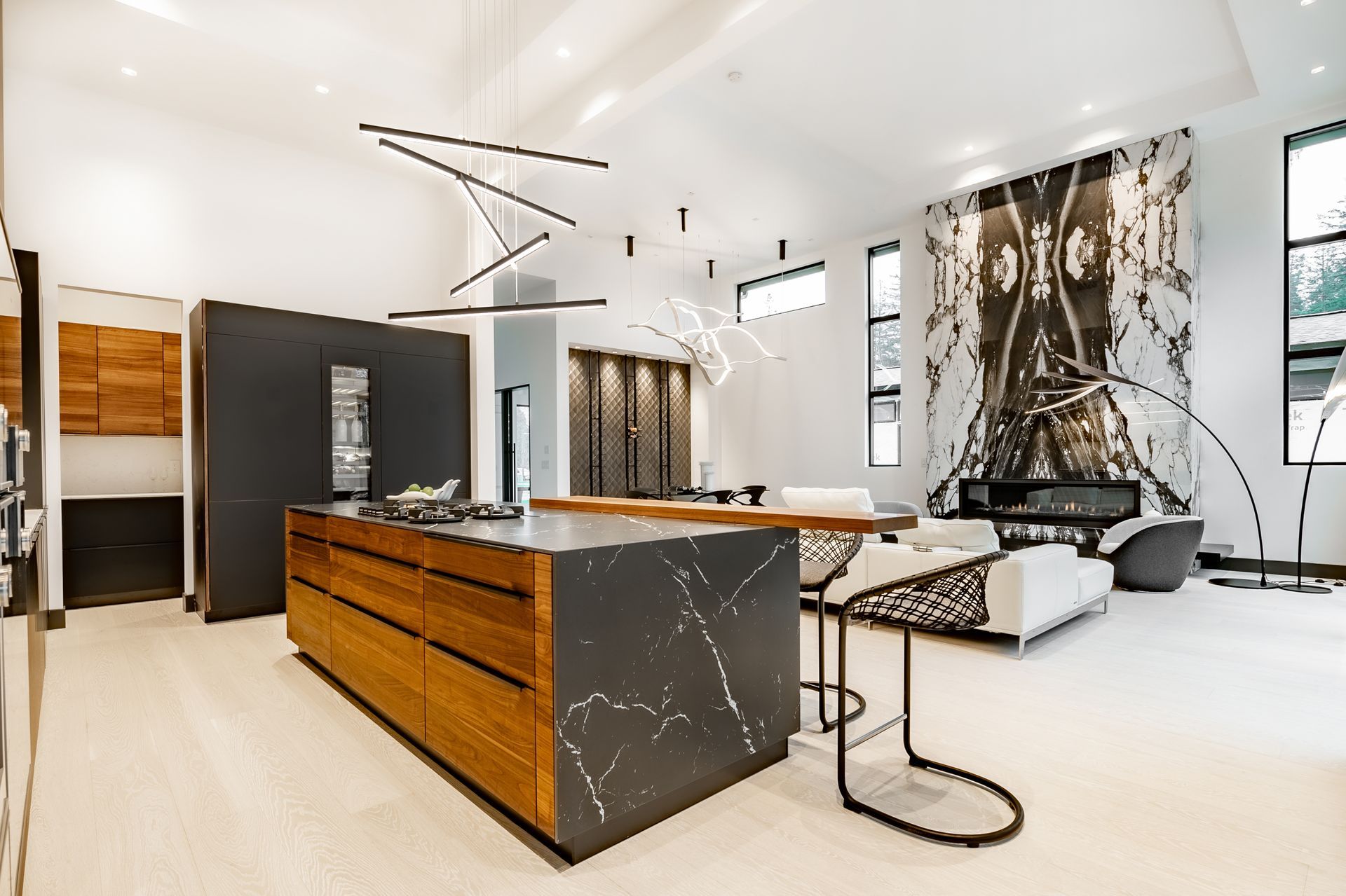 A modern kitchen features a marble island with wood cabinets, high-backed stools, and a dramatic marble fireplace wall.