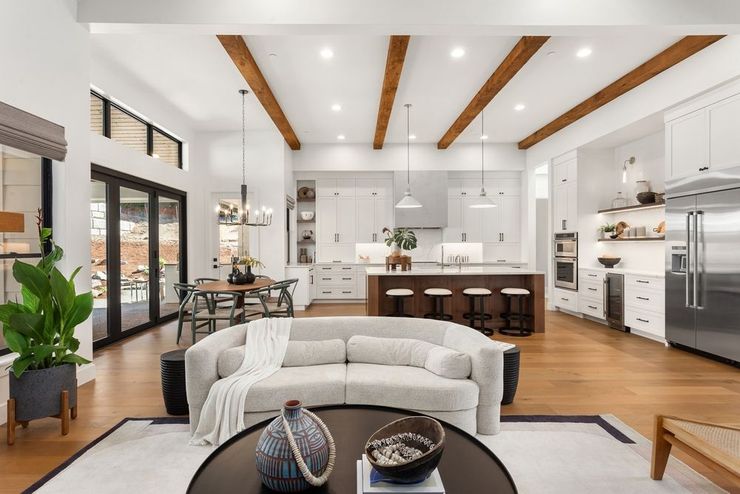 An open-concept living room and kitchen featuring a cream curved sofa, wood ceiling beams, and white cabinetry.