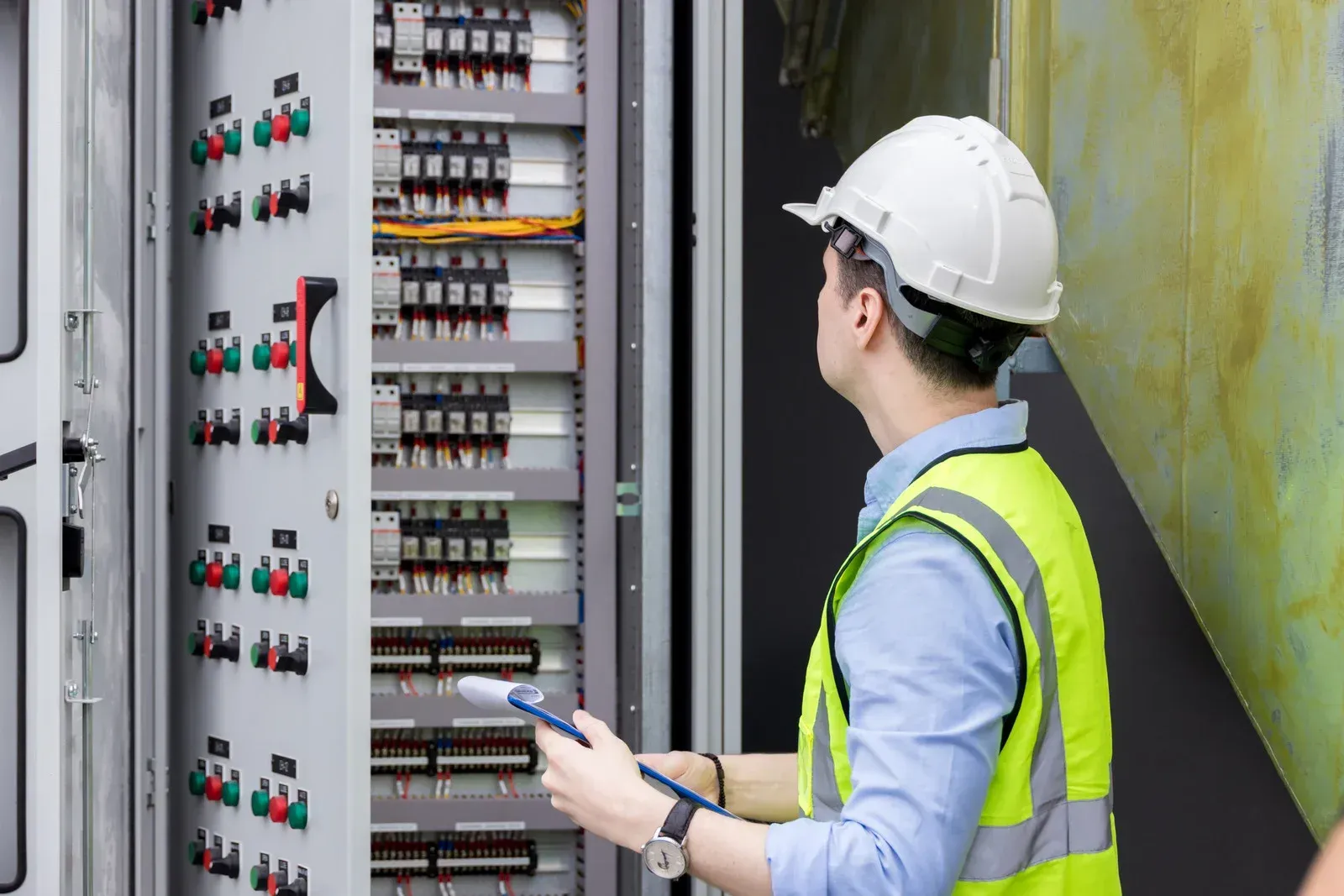 Electrician in hard hat and safety vest inspecting electrical panel with a tool.