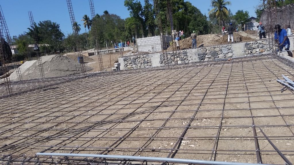 Construction site with a grid of steel rebar on a dirt foundation, surrounded by piles of dirt and stones.