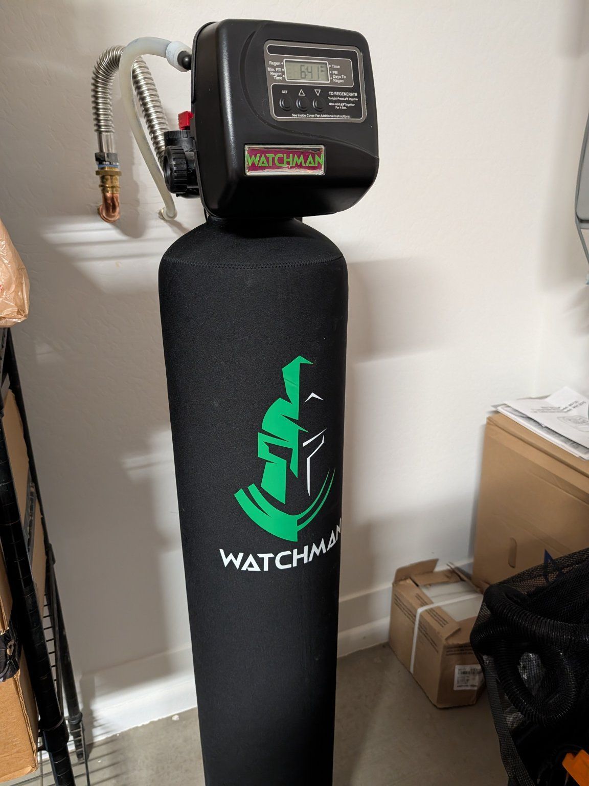 A Watchman water filtration system with a tall black tank featuring a green Spartan helmet logo, standing in a room.