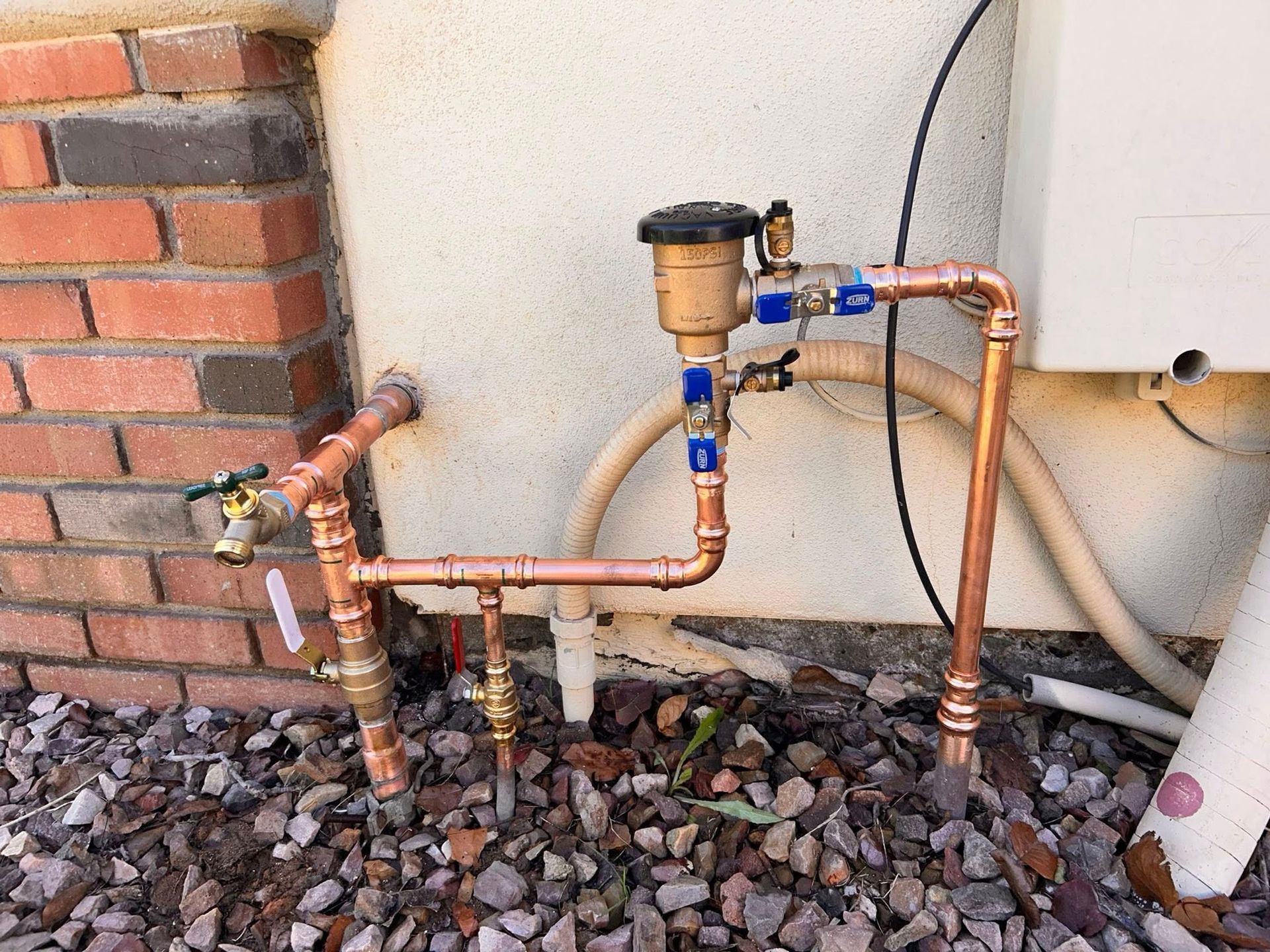 Copper irrigation backflow prevention assembly installed on an exterior stucco wall next to a brick pillar.