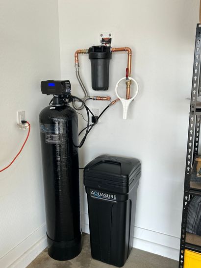 A home water filtration and softening system with a black tank, a filter housing on the wall, and a salt brine container.