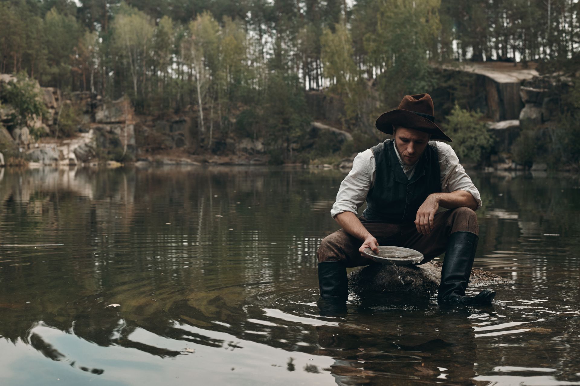 Person in a black hat crouches in a shallow pond, washing clothes with trees reflected in the water.