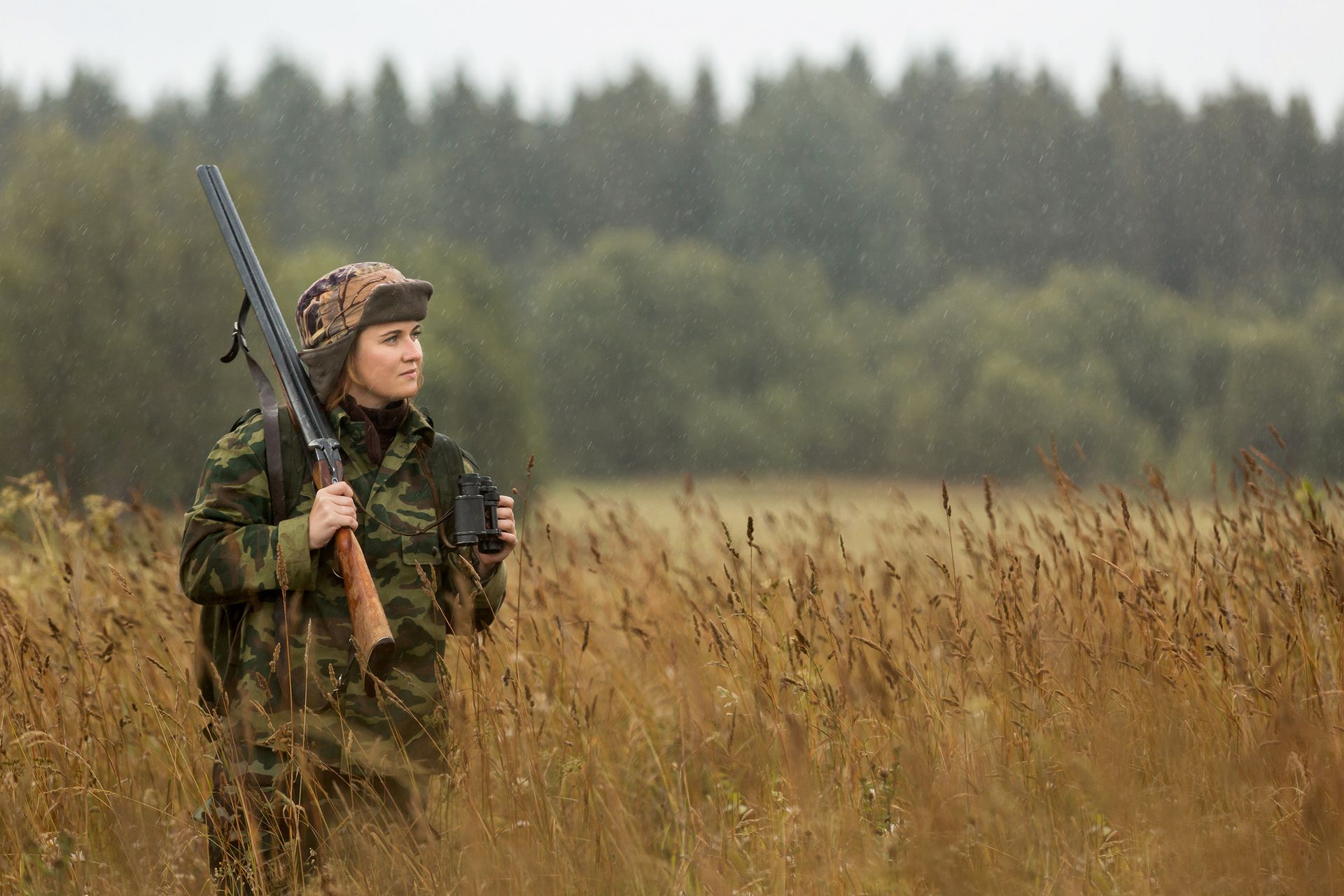 Hunter in camouflage holding a shotgun in a grassy field with a forest in the background