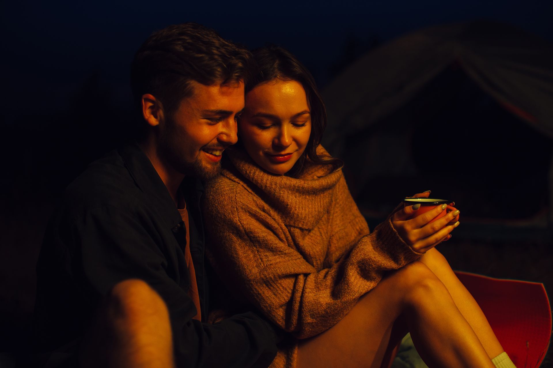 Couple cuddles near campfire, woman holding mug, dark setting.