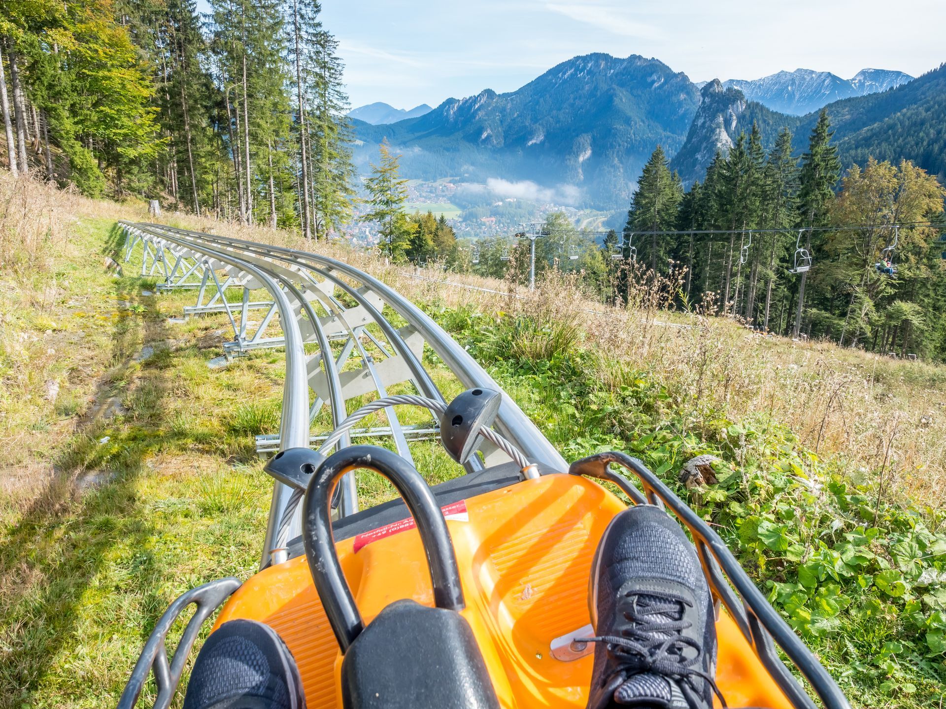 A mountain coaster ride descending through trees, with a rider’s feet visible and snowy peaks ahead.