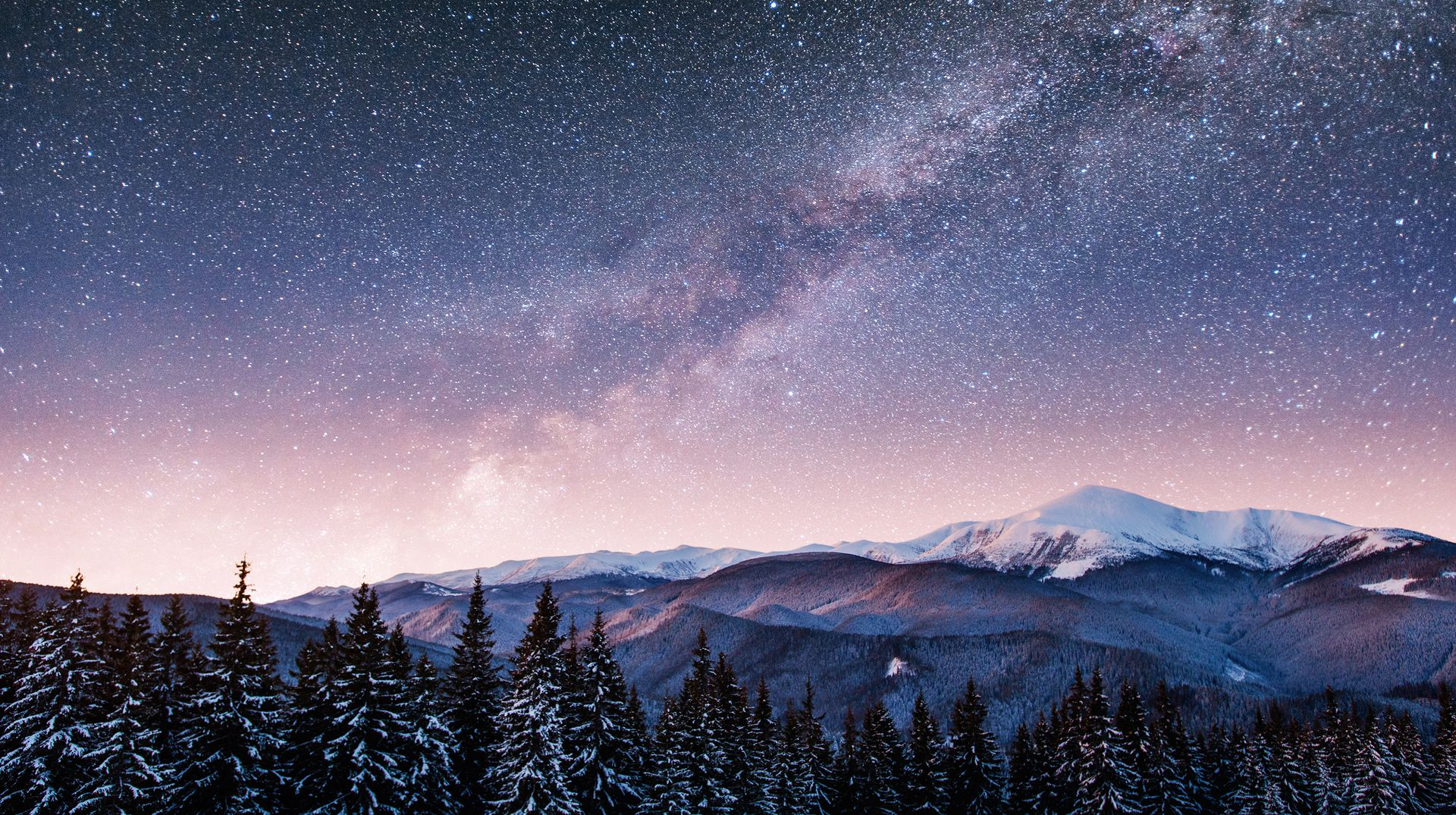 Snow-covered mountains and evergreen trees under a starry night sky with the Milky Way visible.