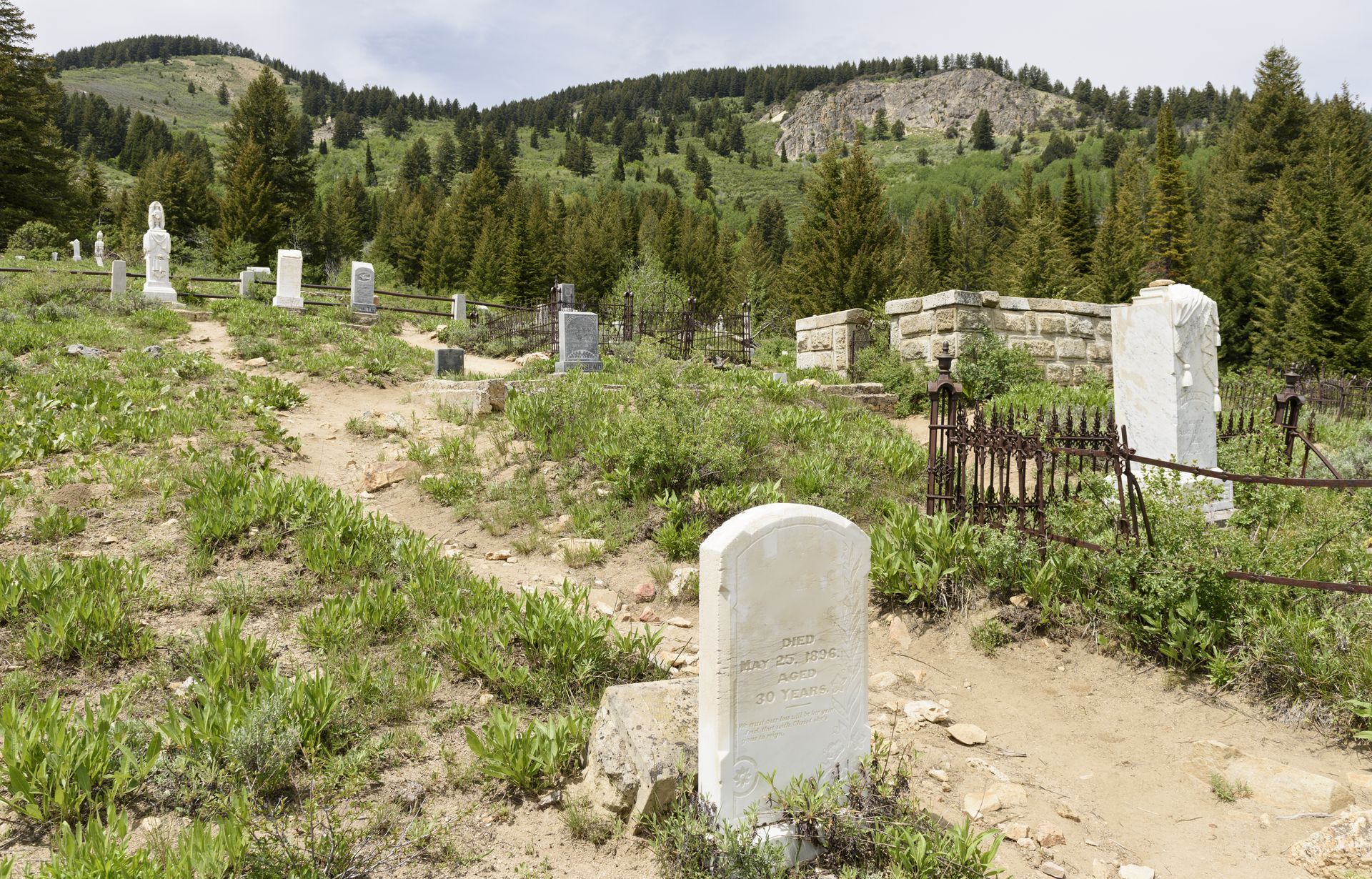 A hillside cemetery with weathered headstones and a dirt path, set against a backdrop of green trees and mountains.