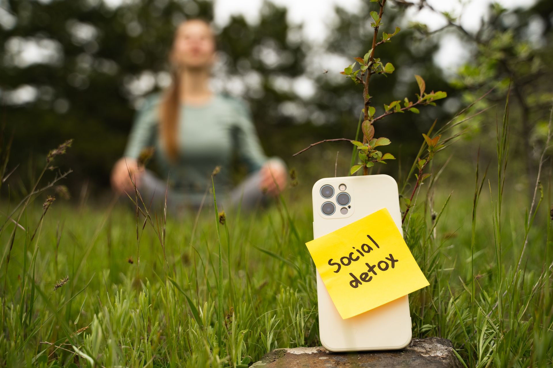 Phone with “social detox” sticky note in grass, with a blurred person meditating in the background