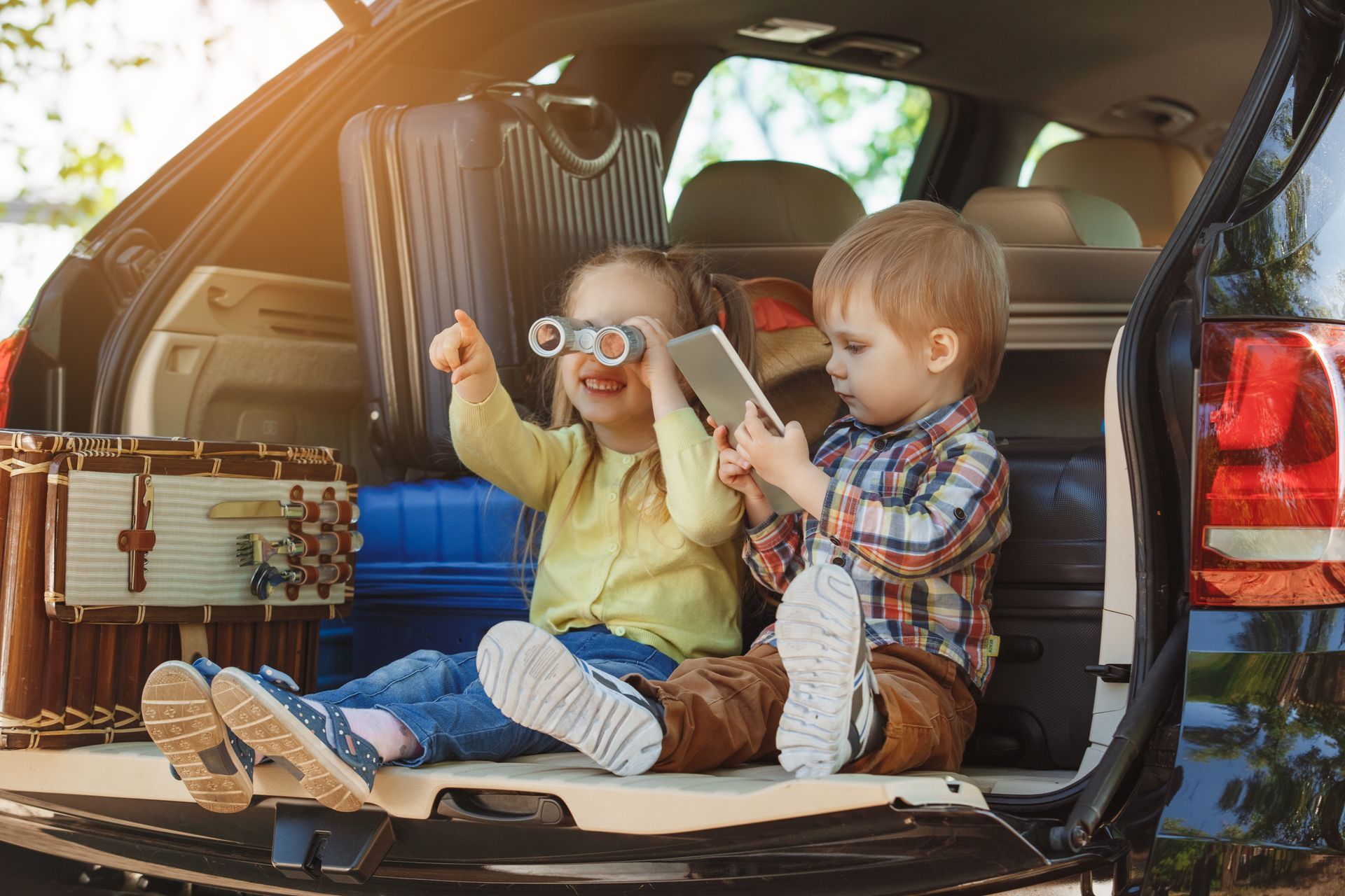 Two children sit in a car trunk packed with luggage; one looks through binoculars while the other uses a tablet.