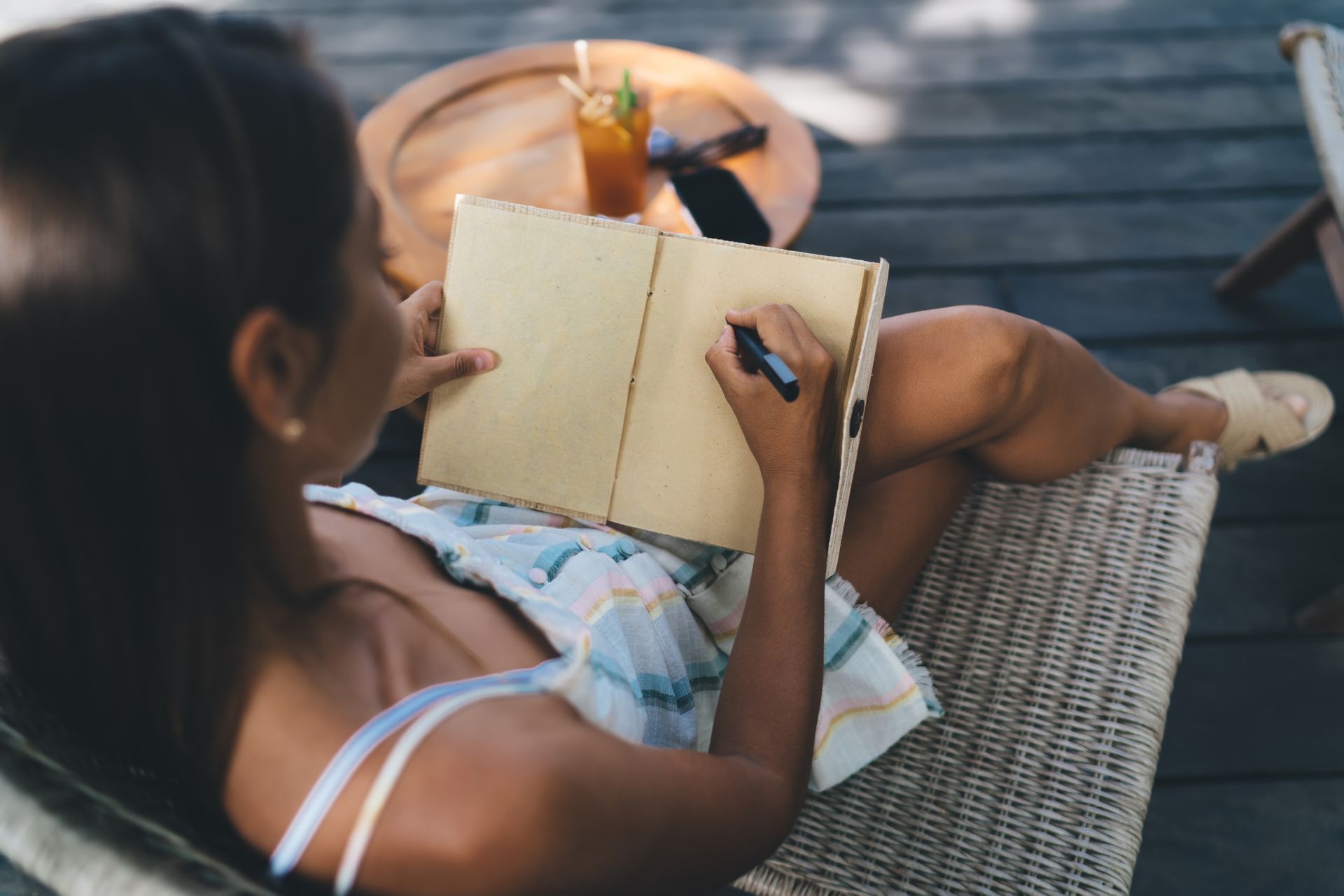 Person lounging on a wicker chair, writing in a notebook beside a round table outdoors