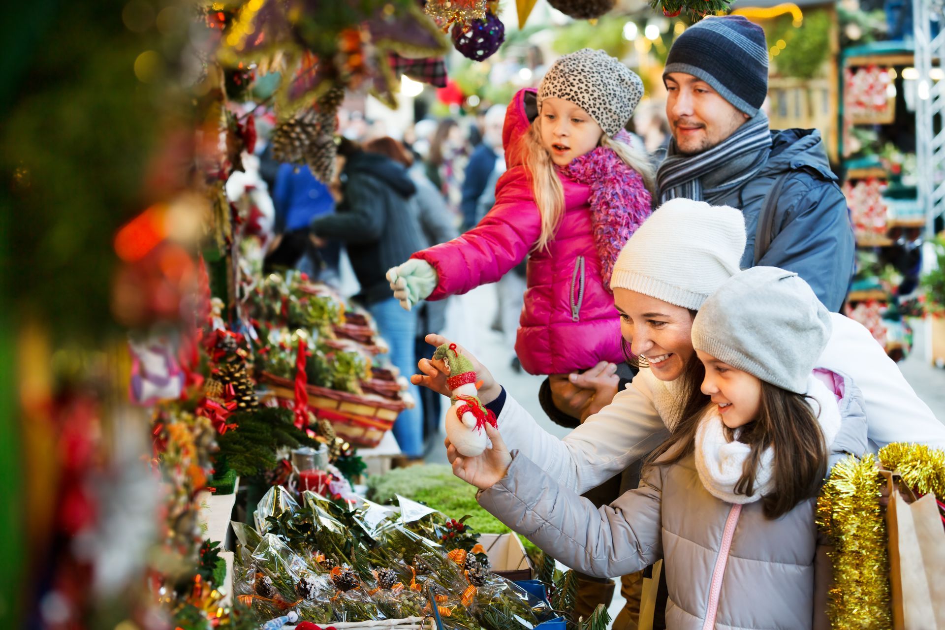 Family at a Christmas market, examining ornaments. Children in winter coats, parents smiling, festive decorations.