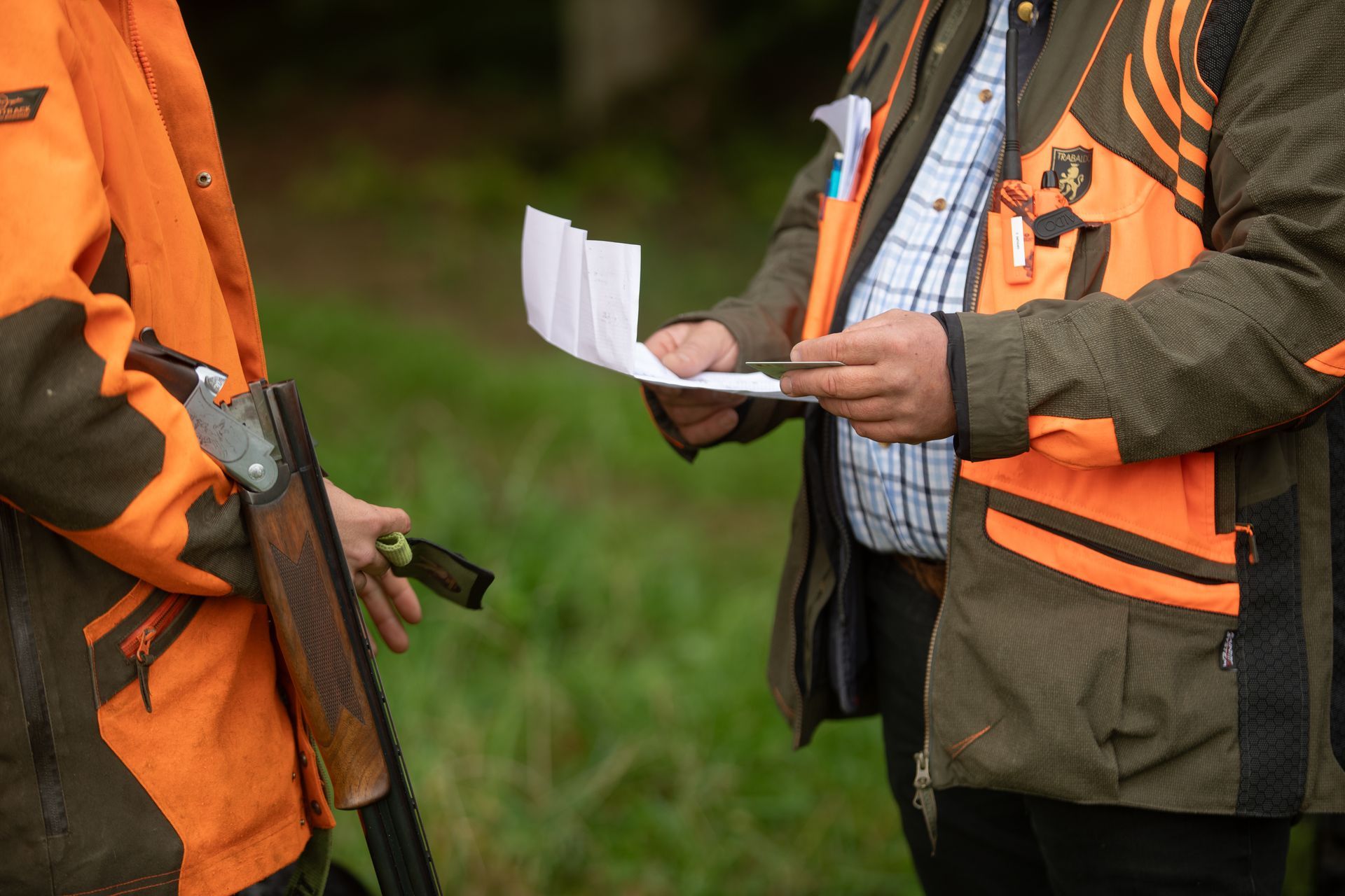 Two hunters in orange vests reviewing papers in a grassy field with a rifle visible