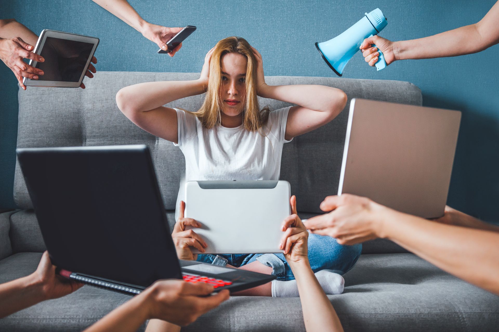 An overwhelmed person on a couch surrounded by hands holding devices and paperwork in a chaotic work scene