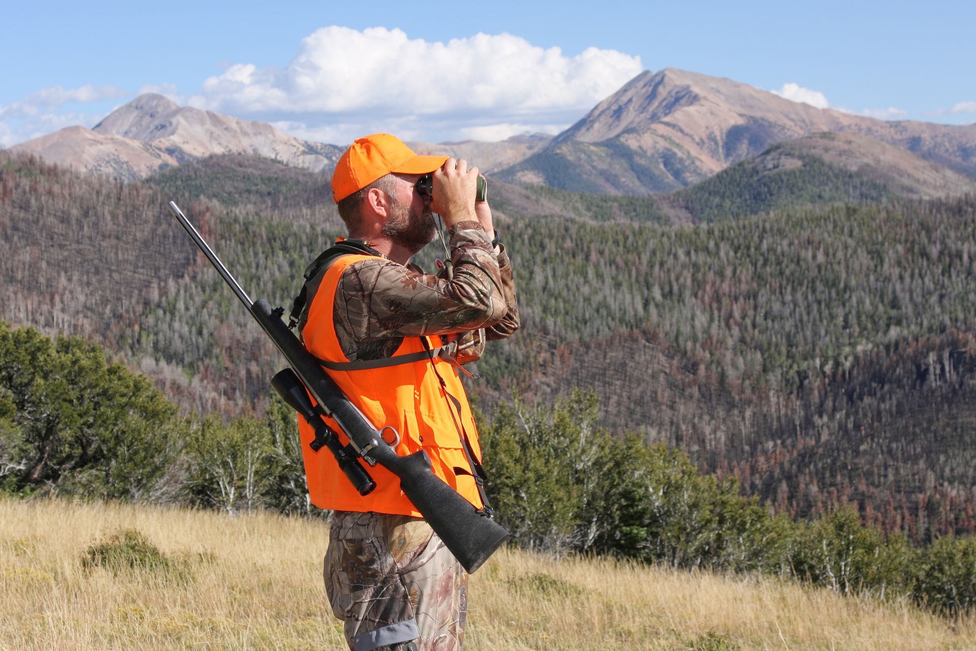 Hunter in orange vest and cap scanning mountain landscape with binoculars, rifle slung over shoulder
