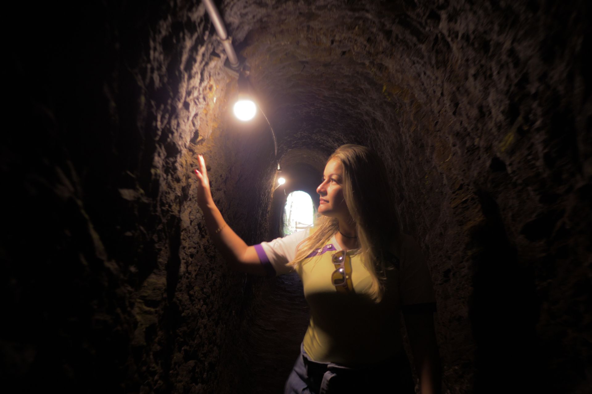 Woman standing in a dark tunnel, reaching toward a glowing light overhead