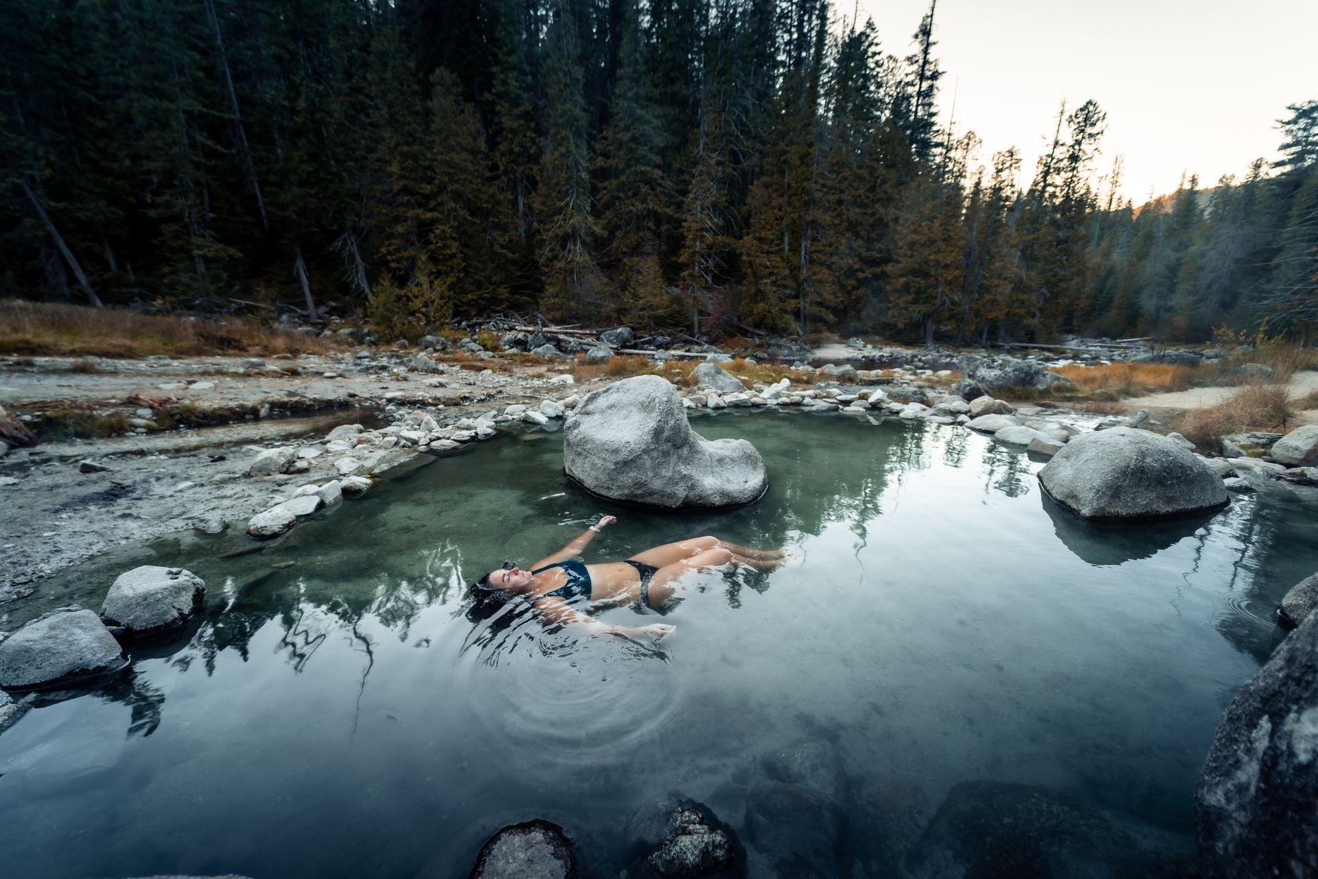 Woman floating in a natural hot spring, surrounded by rocks and evergreen trees.