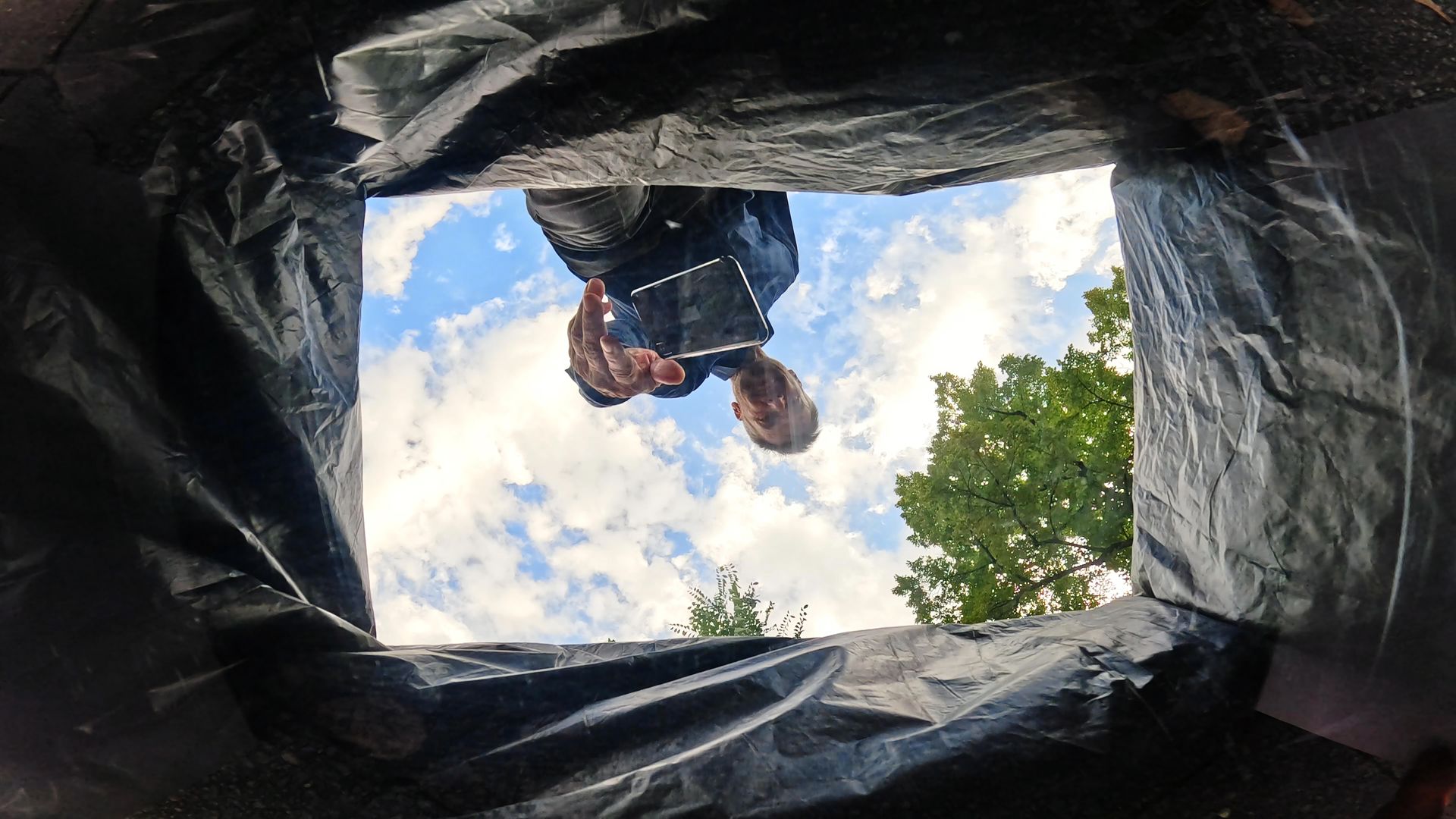 View from inside a tent opening to a cloudy sky, trees, and a person leaning over the entrance.