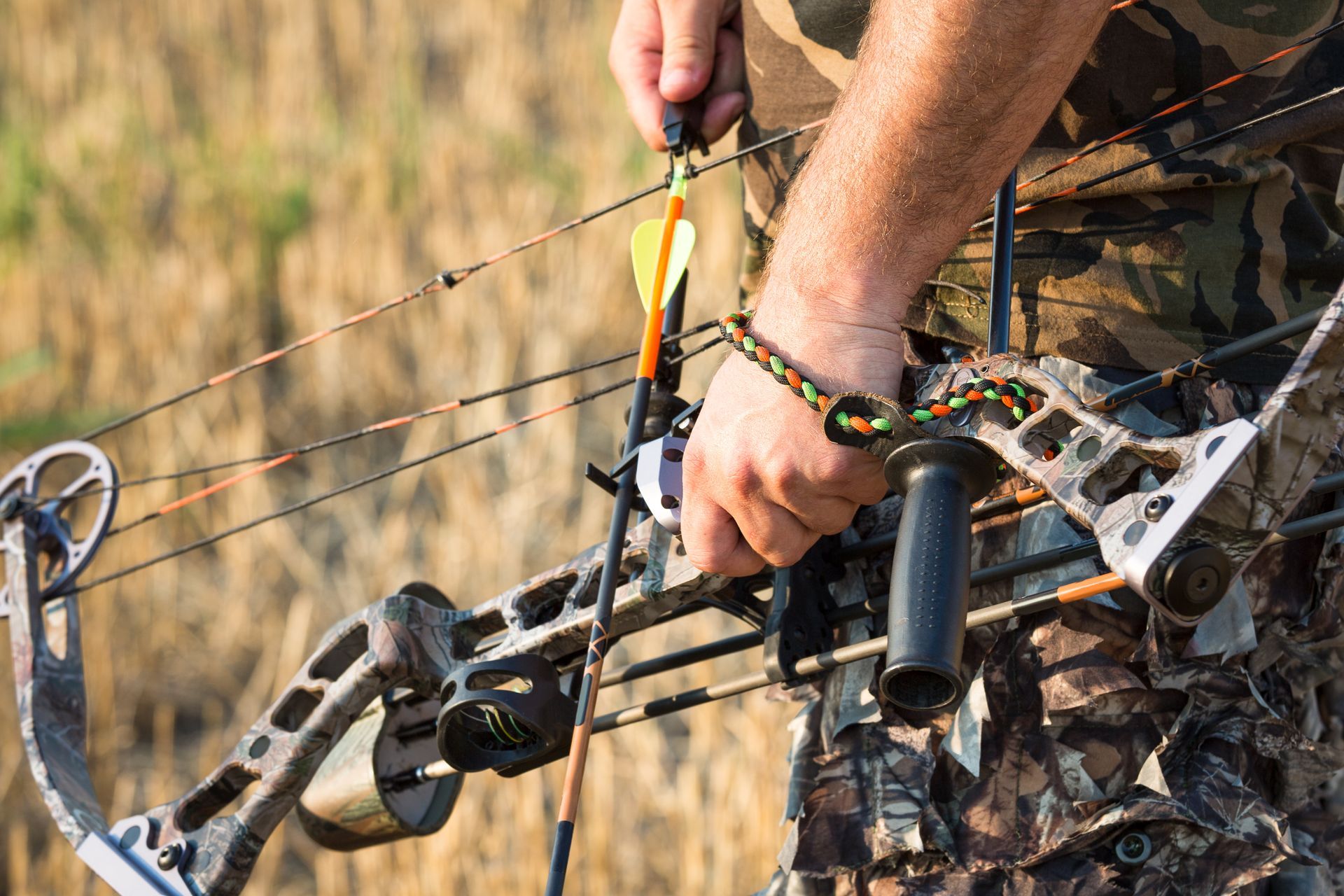 Person holding a compound bow with an arrow, wearing camouflage outdoors near a fence