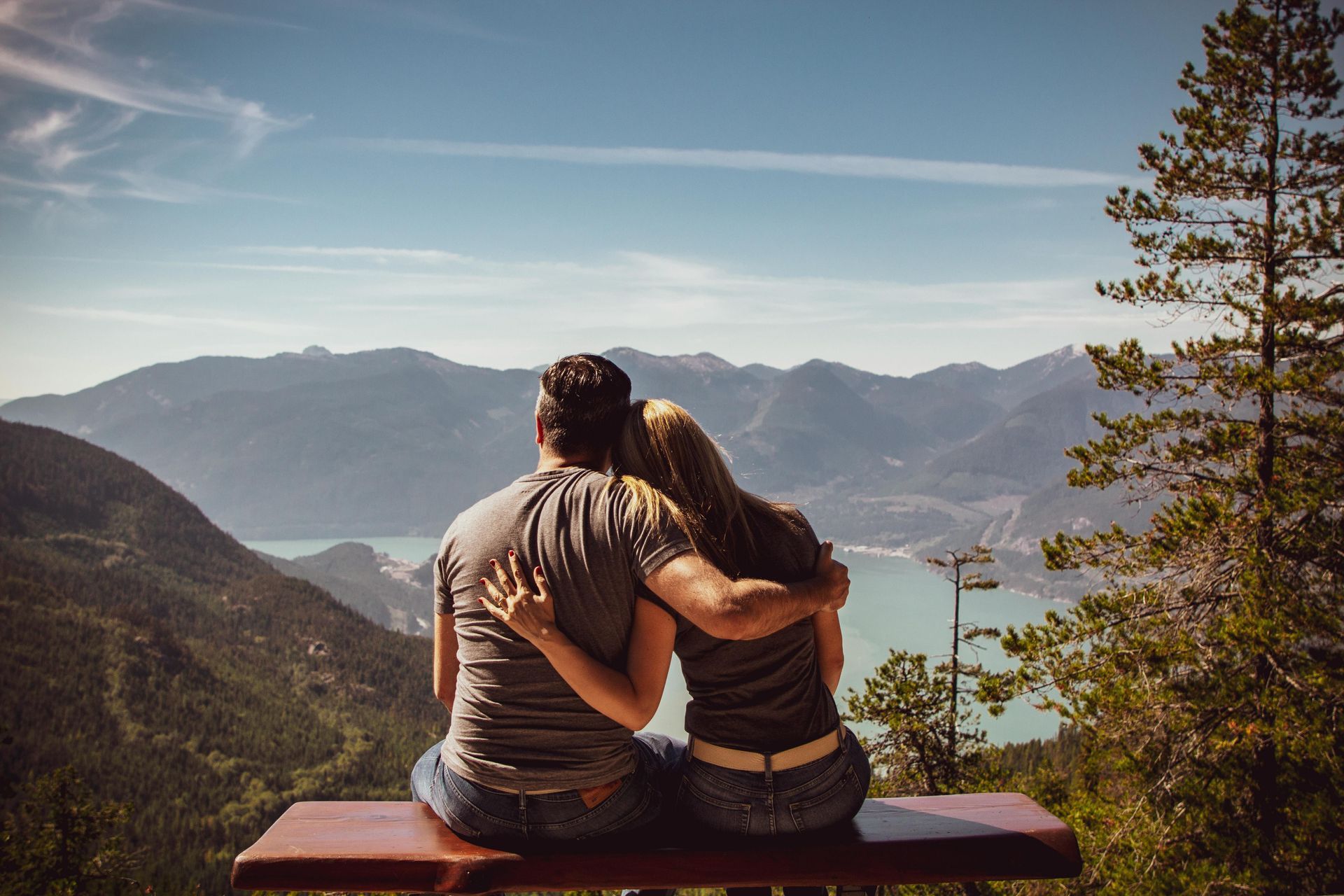 Couple with arms around each other, gazing at mountain landscape over a lake. Blue sky, brown bench.