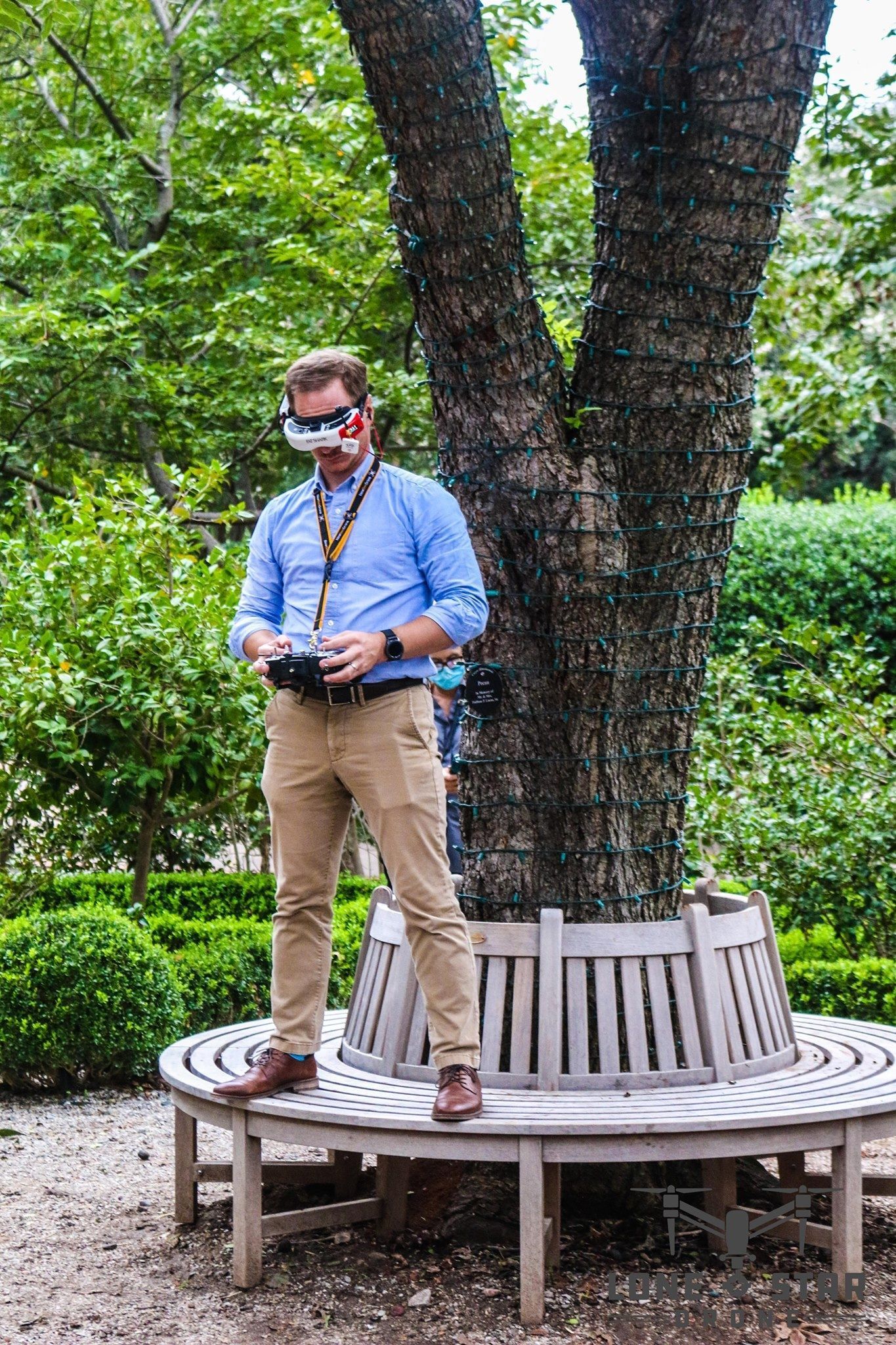 Man in blue shirt and khakis, using drone controller, wears goggles on bench by tree in garden.