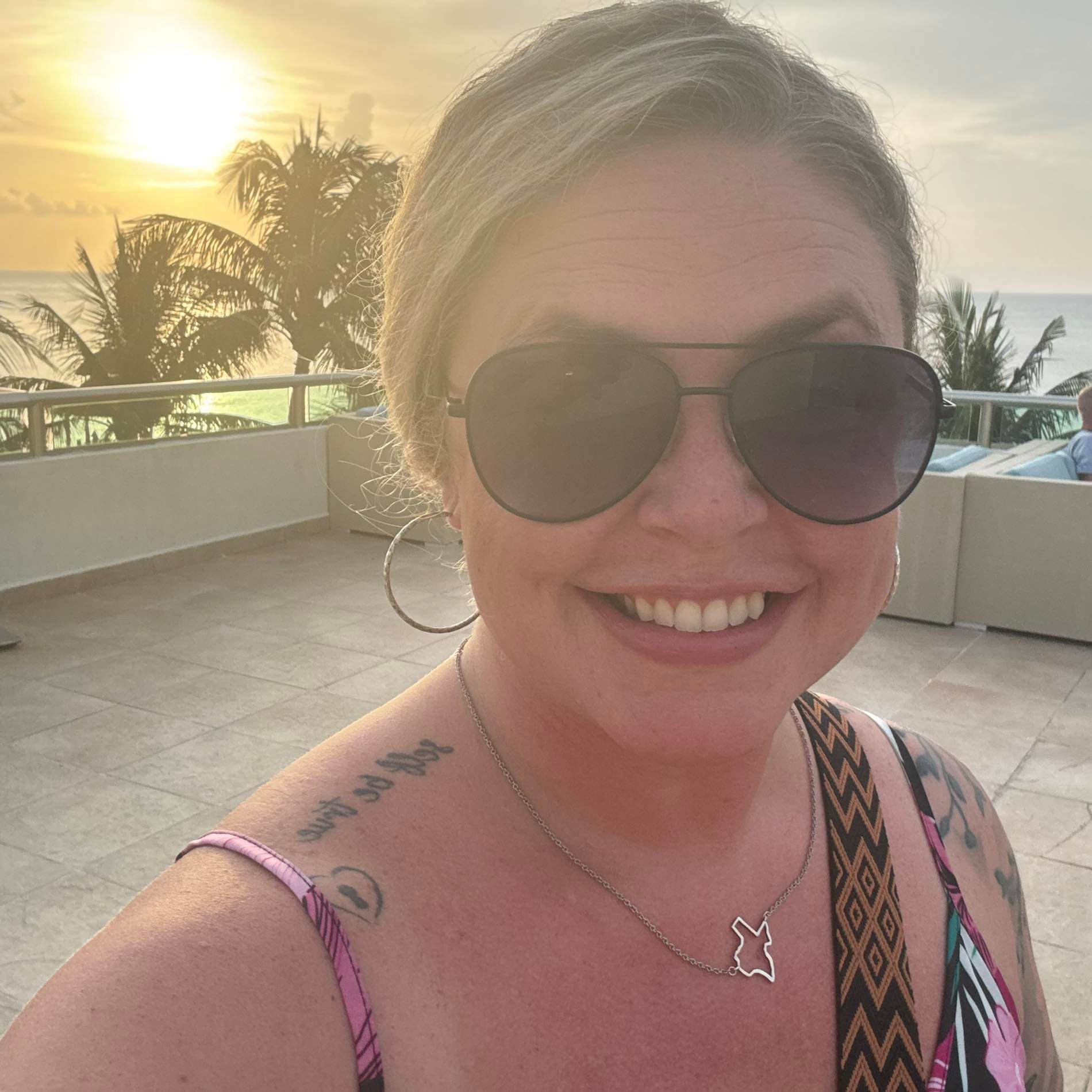 Woman in sunglasses smiles at sunset on a balcony with ocean view.