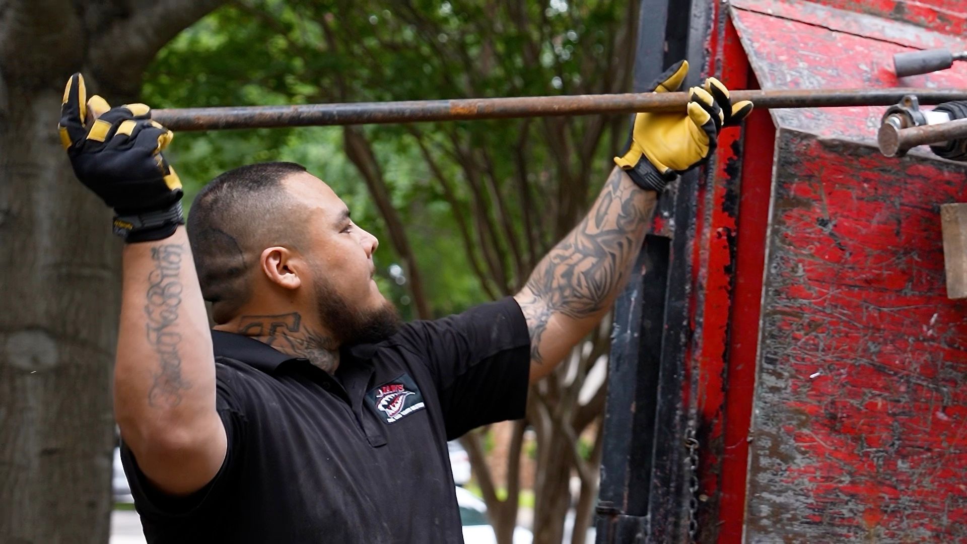 Man with arm tattoos, wearing gloves, raises a metal bar, standing near a red truck.