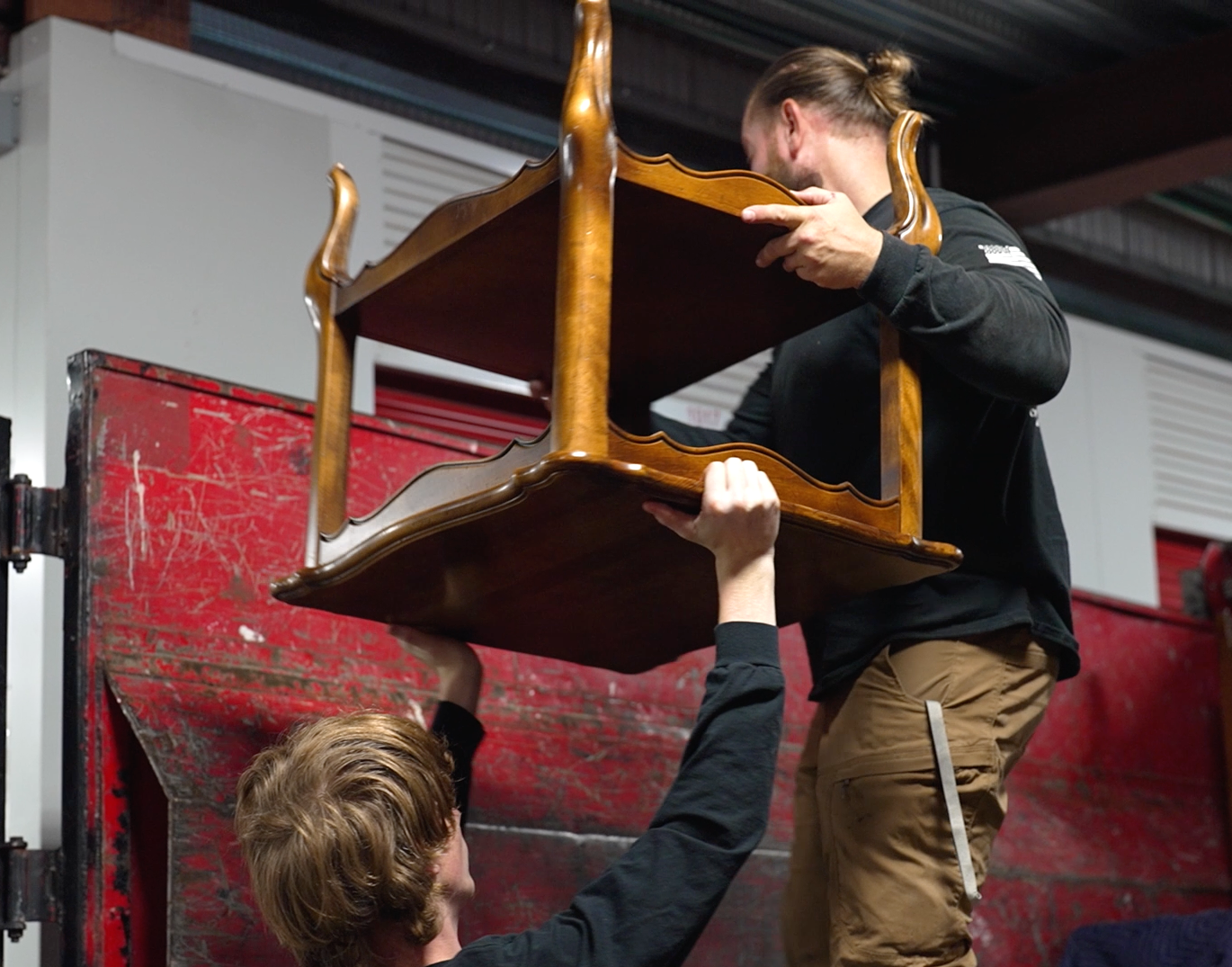 Two men lift a wooden table, placing it into a red dumpster.