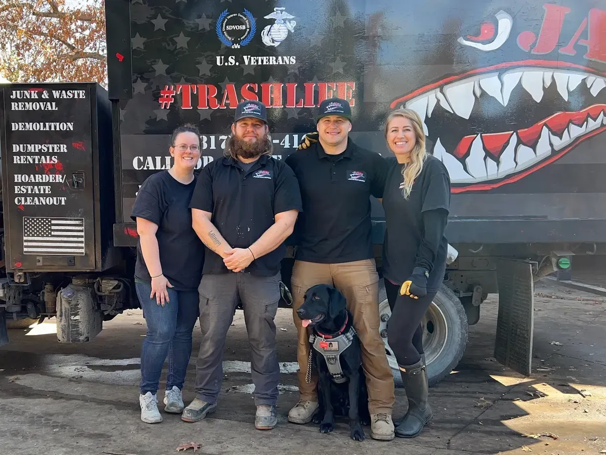 Four people and a dog pose in front of a trash truck. The people are smiling, with the truck in the background.